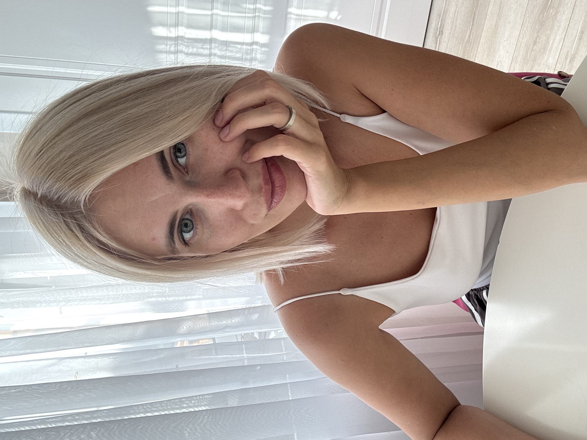 The image shows a woman with blonde hair, a white top, and a ring on her finger. She is leaning forward with her hand on her face, looking at the camera with blue eyes and pink lipstick. She is sitting at a white table, with a window and blinds in the background. The scene appears to be indoors, possibly a home or a studio, with natural light coming from the window. The woman's expression is relaxed and friendly. There are no visible text elements in the image.