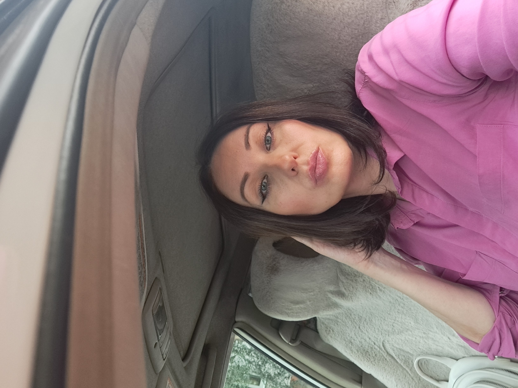A medium shot shows a woman with dark brown hair and fair skin sitting in the driver's seat of a car. She is wearing a bright pink collared shirt and has on winged eyeliner and lip gloss. Her left hand is resting on her head, and her right arm is bent at the elbow. The interior of the car is visible, including a gray headliner and a tan seat with a plush cover. The woman is looking directly at the camera with her lips pursed. The background outside the car window appears to be foliage. The image is a selfie taken from inside the vehicle.