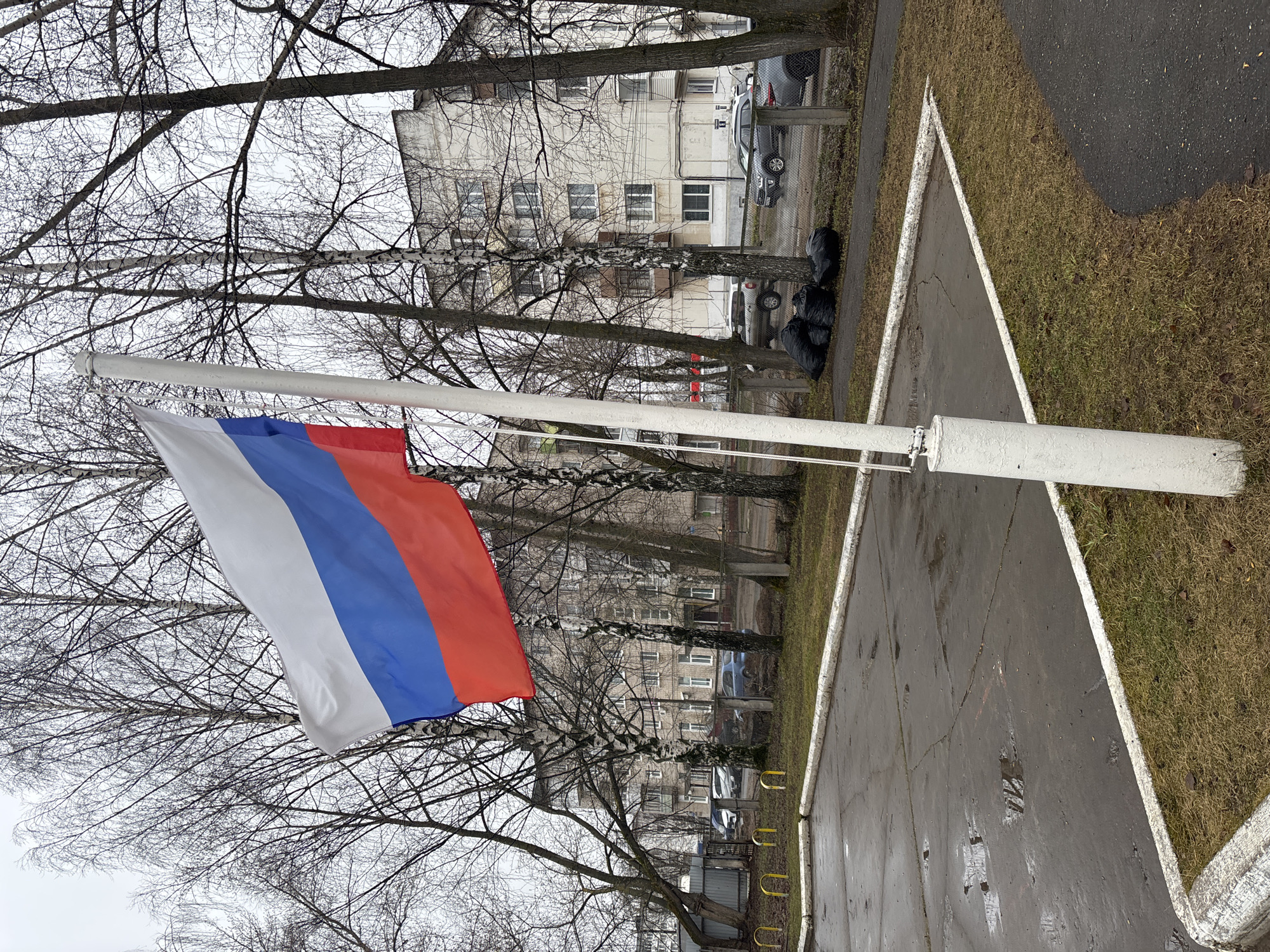 The image shows the flag of Russia hanging from a white pole in an urban environment, potentially in Chaikovsky, Russia. The flag is prominently displayed against a backdrop of bare trees and an apartment building. The ground appears to be a combination of pavement and grass. The scene has a muted, overcast feel. There are parked cars and some black bags visible in the background along a fence line. A painted triangle outlines a section of the pavement. There are no visible people.