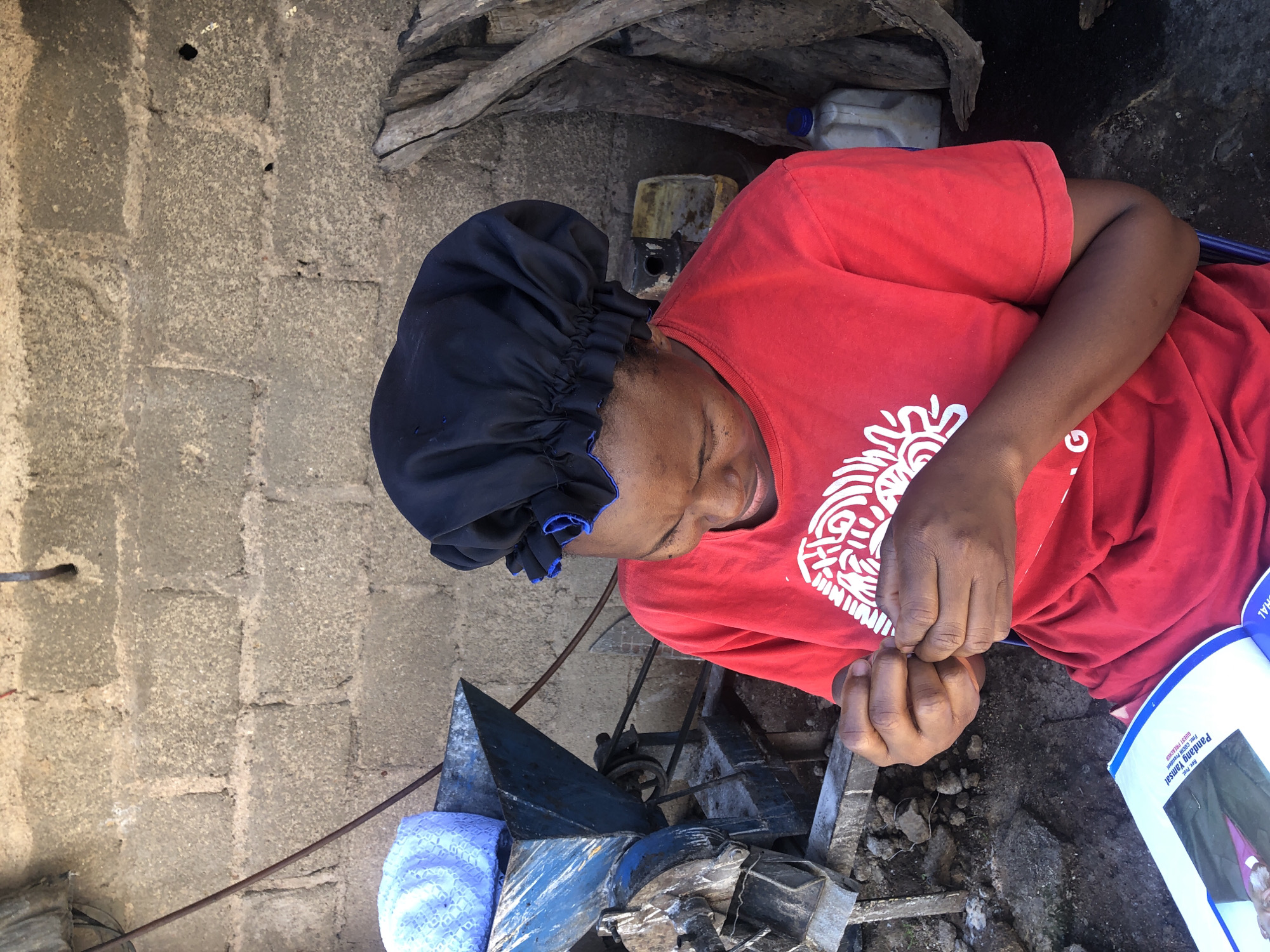A medium shot shows a Black woman, likely in her 20s or 30s, with a black bonnet on her head and wearing a red t-shirt with a white graphic design. She is seated outdoors, looking down and focused on threading a needle with her hands. In front of her is an open book with a picture on the left page. To her left, a blue metal hopper with a white and blue textured object on top sits on a piece of machinery. The background consists of a rough, grey cinder block wall and a pile of dried branches on the right. The scene appears to be an outdoor workshop or processing area, possibly in the city of Jos, Nigeria, given the context.