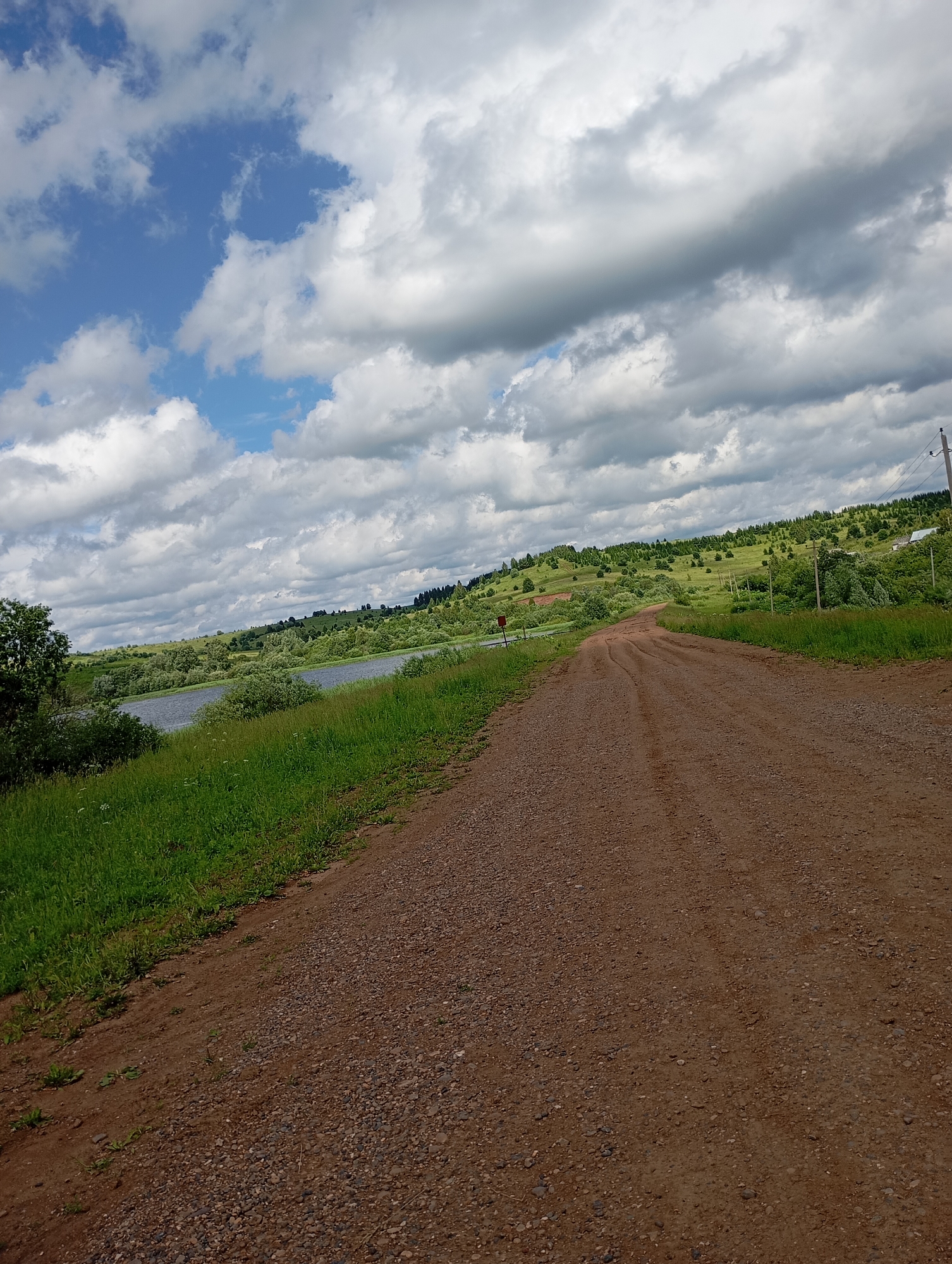 This is a full, eye-level, vertical shot of a dirt road leading through a rural landscape under a cloudy sky. The road is composed of brown dirt and small pebbles, with tire tracks visible. To the left of the road is a grassy embankment leading down to a body of water, likely a lake or wide river. Beyond the water, rolling green hills dotted with trees and occasional small buildings stretch towards the horizon. The sky is filled with dramatic, gray and white cumulus clouds, with patches of blue sky visible. The scene is located in the village of Ryabki, Russia. There are no people or animals present in the image. The environment is natural and rural.