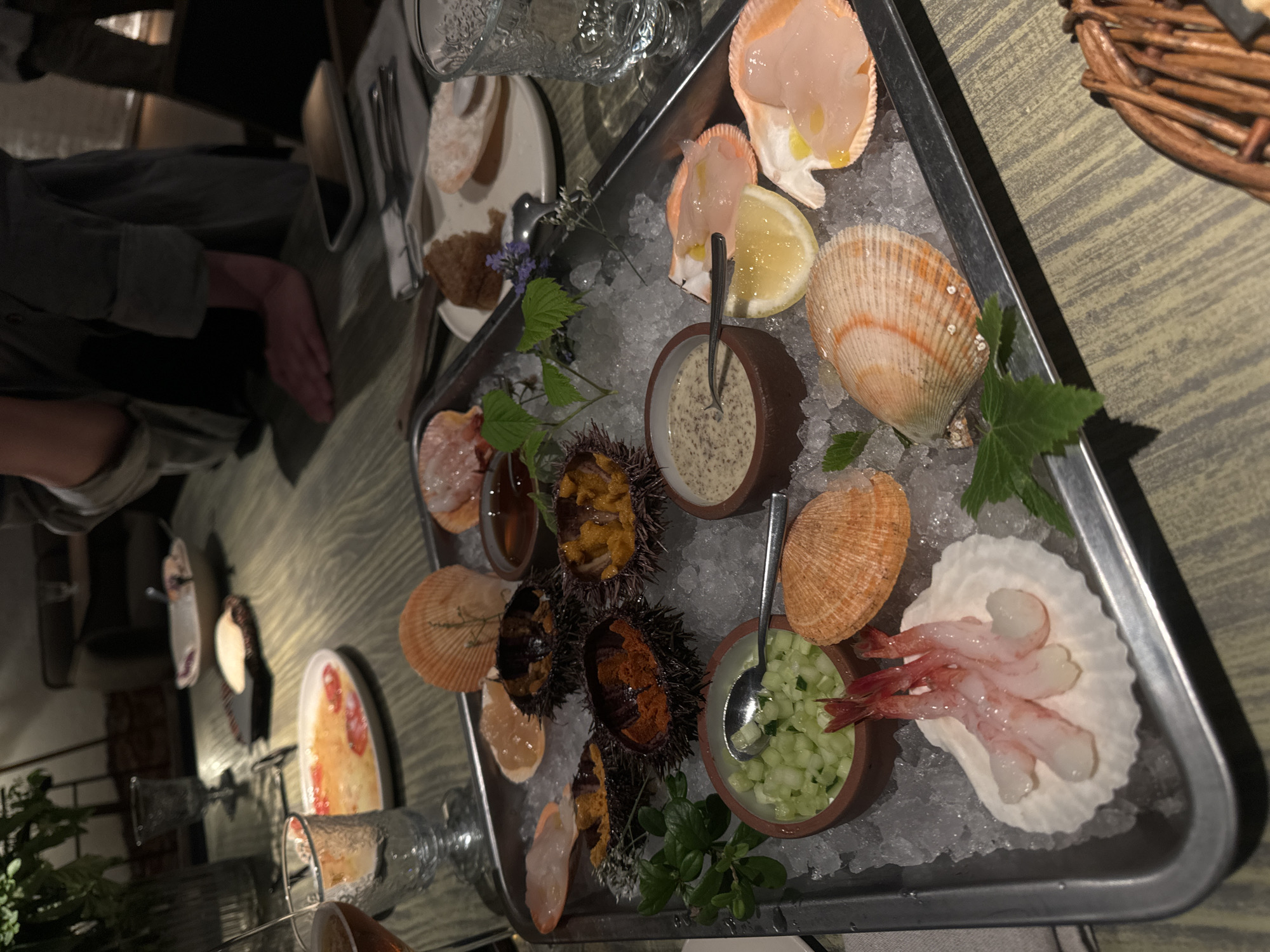 A close-up, eye-level, slightly overhead shot captures a seafood platter served on a metal tray filled with crushed ice. The platter is brimming with various seafood items, including several sea urchins, raw scallops in their shells, a small bowl of chopped cucumber salad with a spoon, a bowl of creamy sauce, and a small dish of sauce. Three raw shrimp are presented on a white shell. Garnishes of green herbs and a sprig of purple flowers add color.
In the background, out of focus, a table setting suggests a restaurant environment. A glass of amber liquid, possibly wine or beer, sits on the left. Another glass, partially obscured, is on the right. Plates with food and a cell phone are also visible on the table. The lighting is dim, typical of a restaurant atmosphere, casting shadows and highlighting the textures of the food. The overall scene suggests an appetizer or a special seafood dish being served. The image does not provide any specific visual cues to indicate its location in Saint Petersburg, Russia.