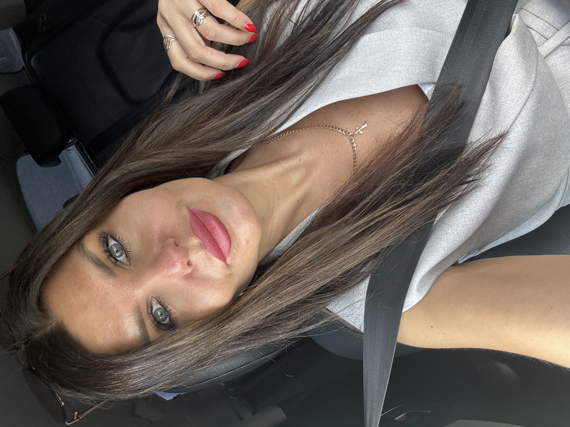 A woman with long, dark brown hair and striking green eyes is captured in a selfie, her head tilted. She is wearing a light grey jacket and a black seatbelt is visible across her chest. A delicate rose gold necklace with a small cross pendant rests on her collarbone. On her left hand, she sports two rose gold rings. Her fingernails are painted a vibrant red. She is wearing rosy pink lipstick, which accentuates her full lips. The background appears to be the interior of a car, suggesting she is a passenger or driver. The lighting suggests it is daytime. There is no visible text in the image.
