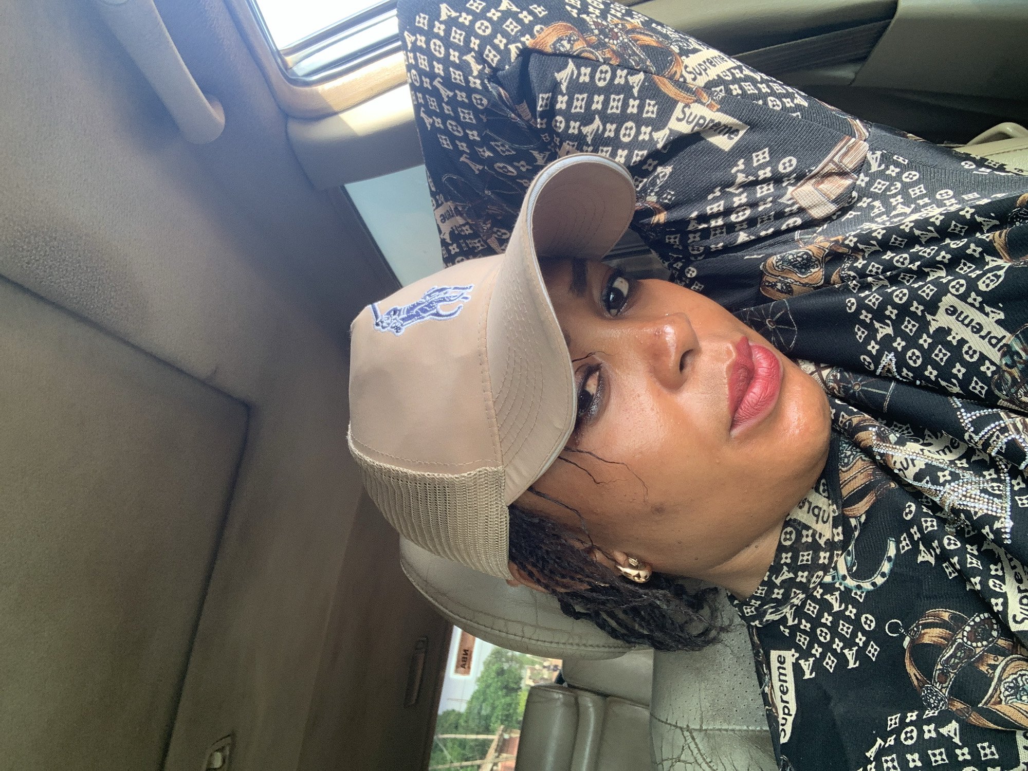 A person is depicted in an upward-angled close-up, appearing to be a selfie taken from inside a car. The individual, who has dark skin, looks directly at the camera with a neutral expression. They are wearing a beige baseball cap adorned with a blue embroidered logo that resembles the Ralph Lauren polo player. Their hair is styled in braids, visible beneath the cap, and they wear dark lipstick and small gold earrings.
The subject is dressed in a long-sleeved black shirt featuring a repeating pattern that includes the "LV" monogram associated with Louis Vuitton, alongside the word "Supreme" in white rectangular boxes. The pattern also incorporates illustrations of ornate belts, buckles, and chains.
The setting is the interior of a vehicle, with a light-colored headliner, a grab handle, and a sunroof visible overhead, suggesting bright daytime lighting. The car's seats, which appear to be light-colored and show signs of wear, are partially visible in the lower part of the frame. Through the car's windows, glimpses of an outdoor environment are seen, including greenery and indistinct structures, indicative of an urban or suburban area. The image captures a moment inside a car, likely in Sabon Tasha, Nigeria.