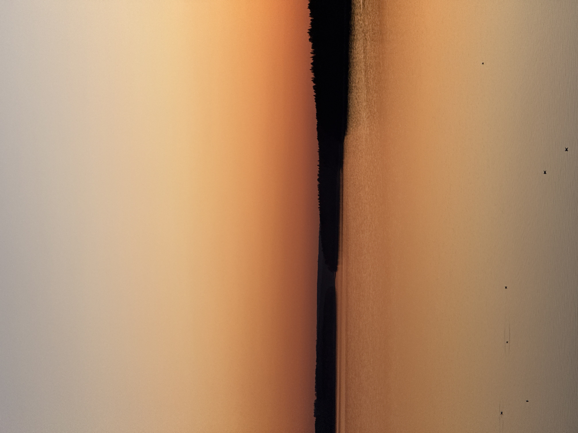 This image shows an aerial view of a body of water, likely a lake or river, with the far shore visible as a dark line dividing the water from a reddish-orange expanse. The water reflects a similar orange hue, potentially from the sky at sunset or sunrise. There are several small, dark objects visible on the water surface, possibly small boats or markers. The setting could be a rural area, as indicated by the presence of a large body of water and the absence of explicit urban structures. Given that the request includes Votkinsk, Russia, it is likely that this is the Votkinsk Reservoir.