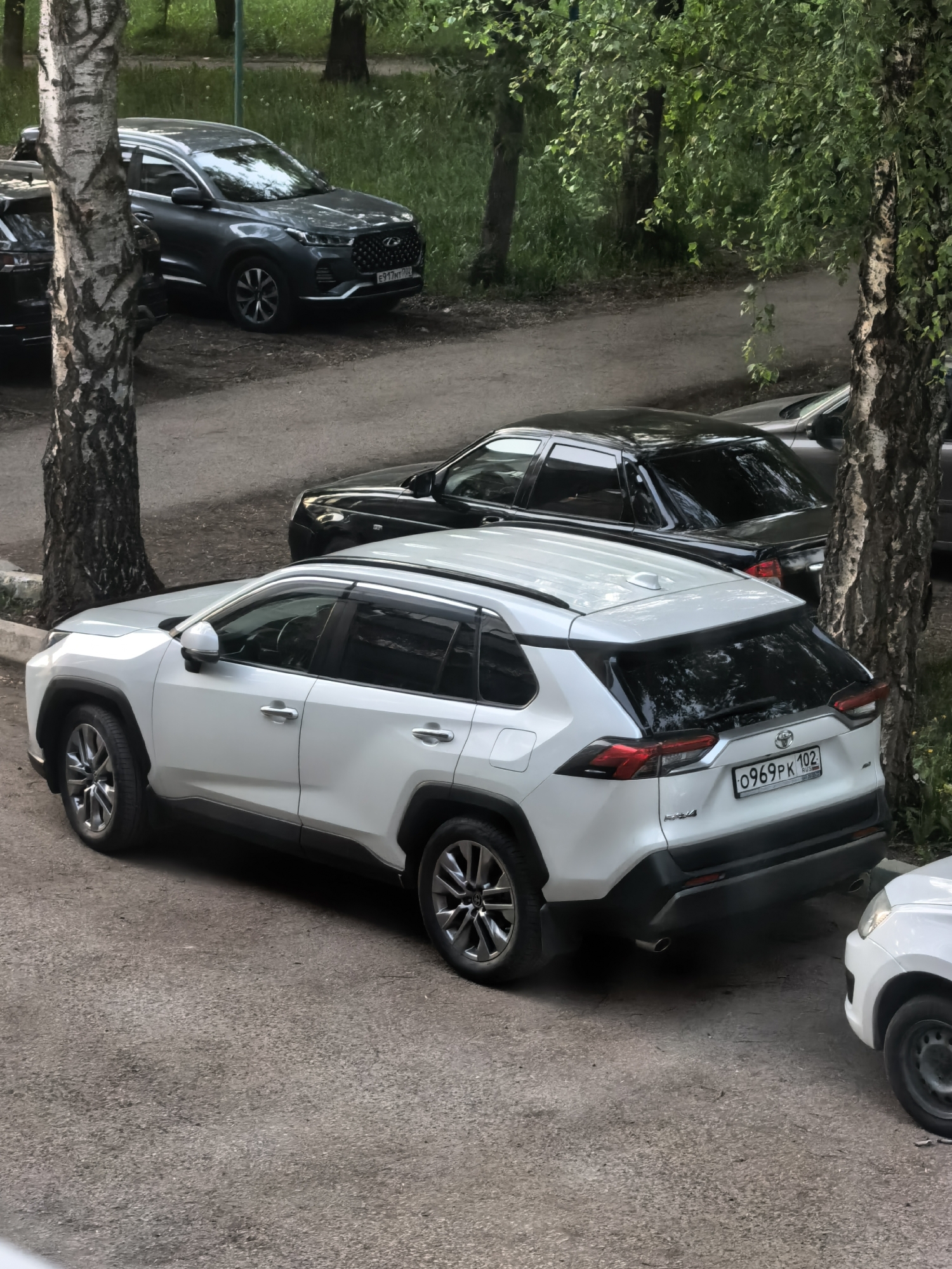 The image depicts a street scene in Sterlitamak, Russia. Visible are several parked cars including a white Toyota RAV4 with license plate "O969PK 102," a black sedan, and a gray SUV. Trees are present in the background and alongside the parked cars. The road surface is asphalt. There are no people visible in the image.