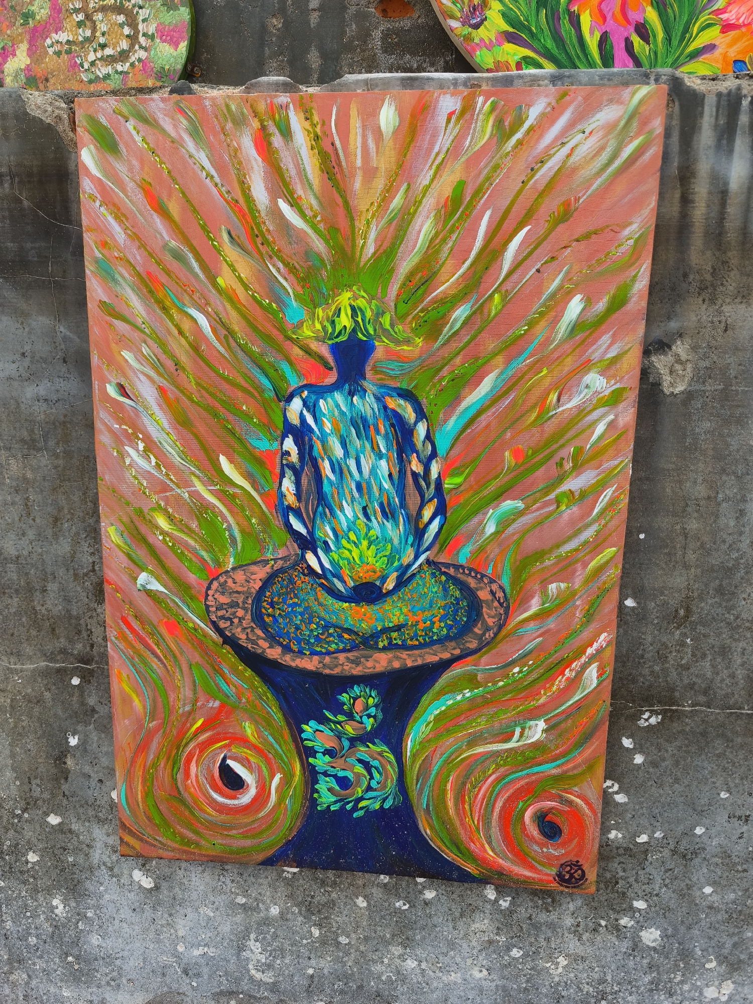 The image shows a rectangular painting of a figure in meditation, rendered in vibrant, abstract brushstrokes. The figure, depicted from the back, is seated on a patterned cushion, with its back adorned in shades of blue and white, and its head topped with a green, spiky crown. The background is a soft orange, filled with numerous white, green, and orange streaks, radiating outwards from the figure. The cushion and the base it rests on are painted in blues and oranges with swirling patterns. The bottom corners of the painting feature large, circular, swirling patterns in red, orange, and green. The painting is leaning against a textured, grey concrete wall, which shows signs of weathering and cracks. To the upper left of the central painting, a portion of another circular painting, featuring colorful floral motifs, is visible. The overall scene suggests an outdoor setting, possibly a wall in the city of Vĩnh Lương, Vietnam, where such artworks might be displayed.