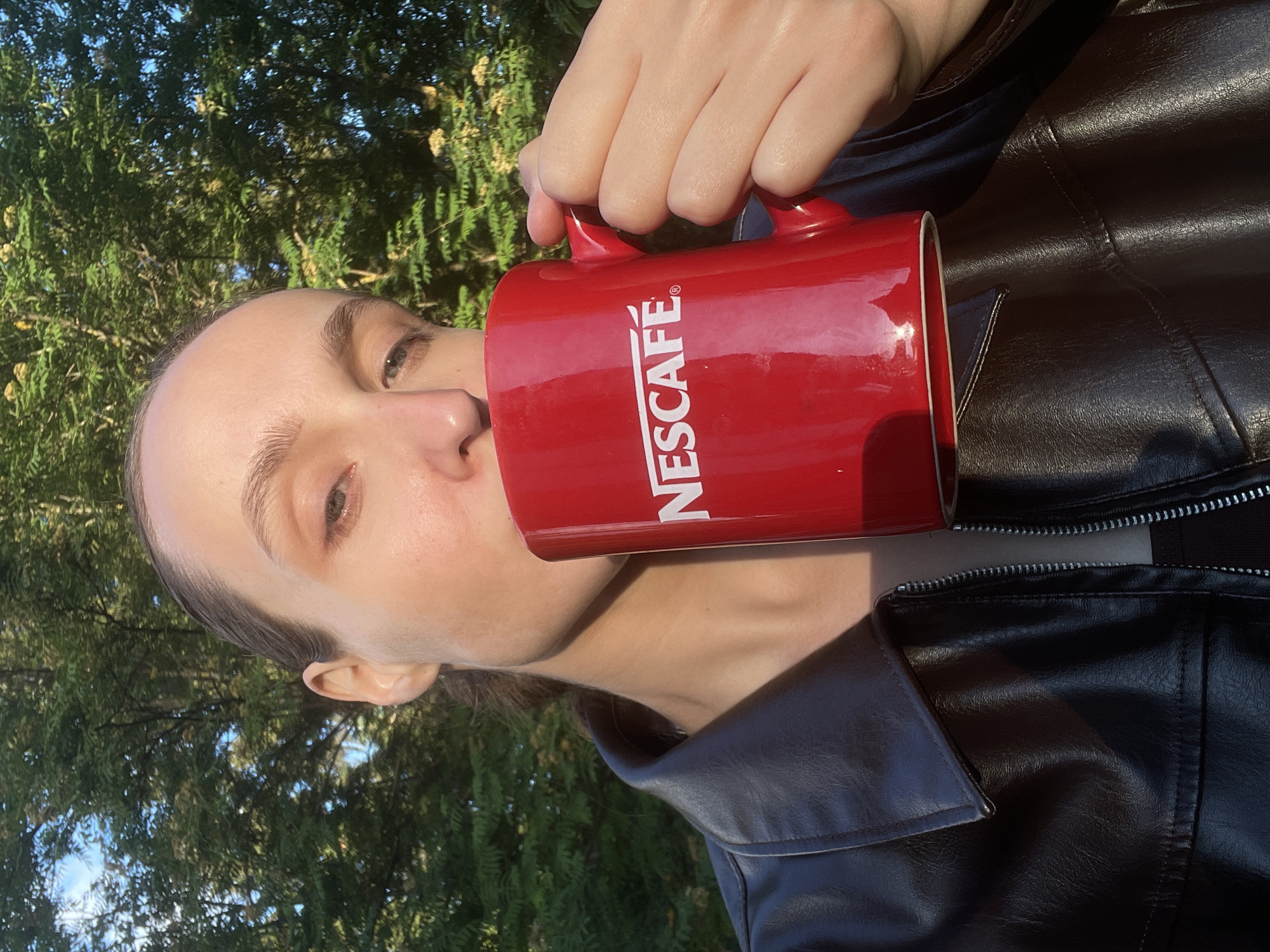 An outdoor, brightly lit close-up photograph captures a person drinking from a red Nescafé mug. The image is rotated 90 degrees clockwise, presenting the individual's head tilted to the left as they bring the mug to their lips. The person, with fair skin and dark eyebrows, is wearing a dark, possibly leather, jacket, visible on their shoulders and upper body. Their right hand holds the handle of the mug, which is made of red ceramic and prominently features the "NESCAFÉ" logo in white, vertical capital letters on its side. The background is composed of lush green trees and foliage, with glimpses of a clear blue sky through the leaves, indicating a sunny daytime setting. Bright sunlight casts highlights across the person's skin, the shiny surface of the mug, and the texture of their jacket. The scene suggests a casual moment enjoying a beverage outdoors in Sharkan, Russia.