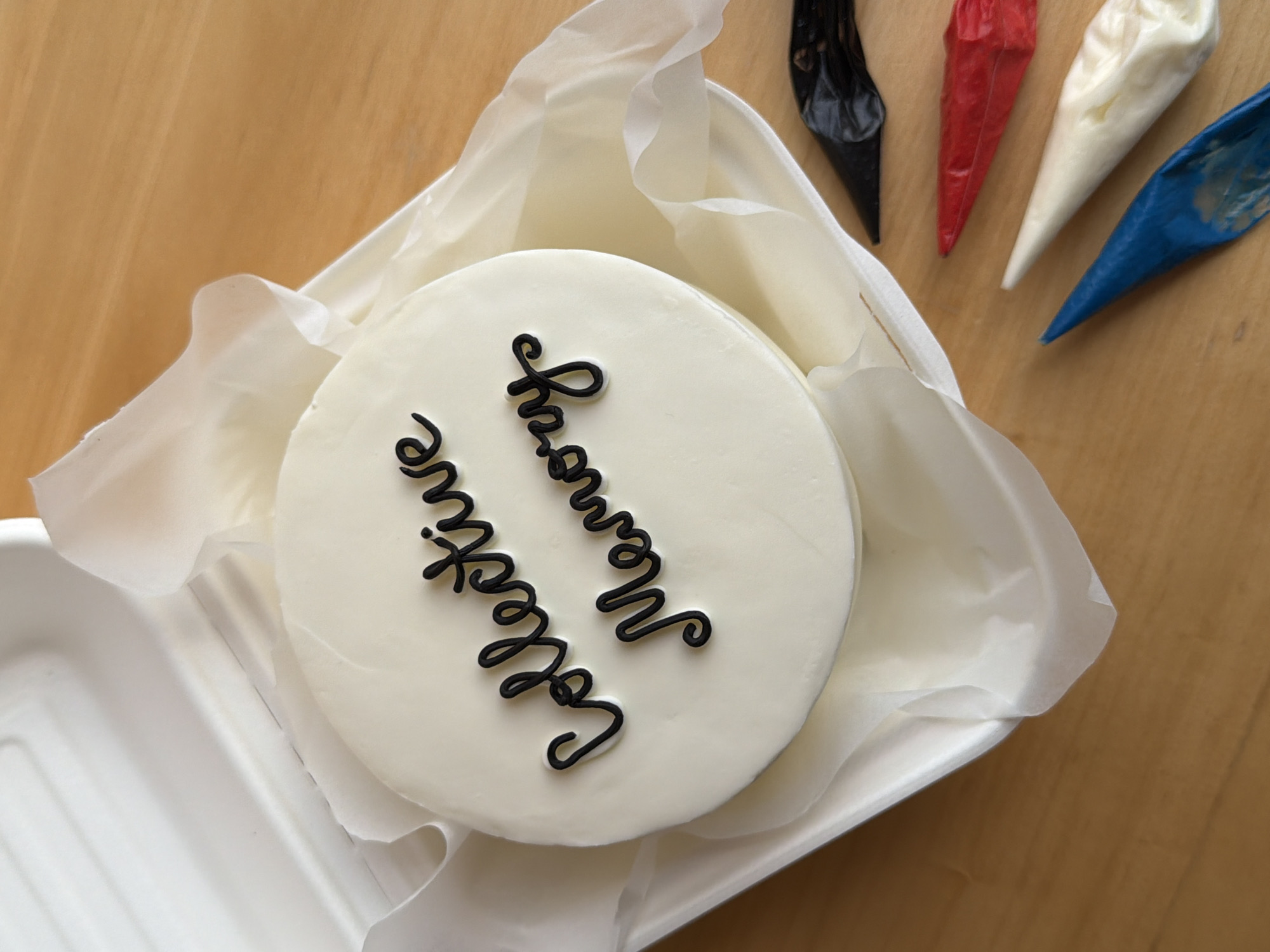 The scene displays a circular cake with white frosting, inscribed with the words "Collective Memory" in black script icing. This cake is contained within a white, rectangular, disposable takeout container, lined with translucent white parchment paper, and resting on a light brown wooden surface. The container's lid is partially visible on the upper left. To the lower right of the container, four small, filled piping bags are arranged on the wooden surface; these appear to contain icing or frosting in black, red, white, and blue. The blue piping bag exhibits some residue near its tip. The wooden surface displays a visible grain pattern. No individuals are present, and no actions are depicted. The described location is in the city of Tel-Aviv, Israel.