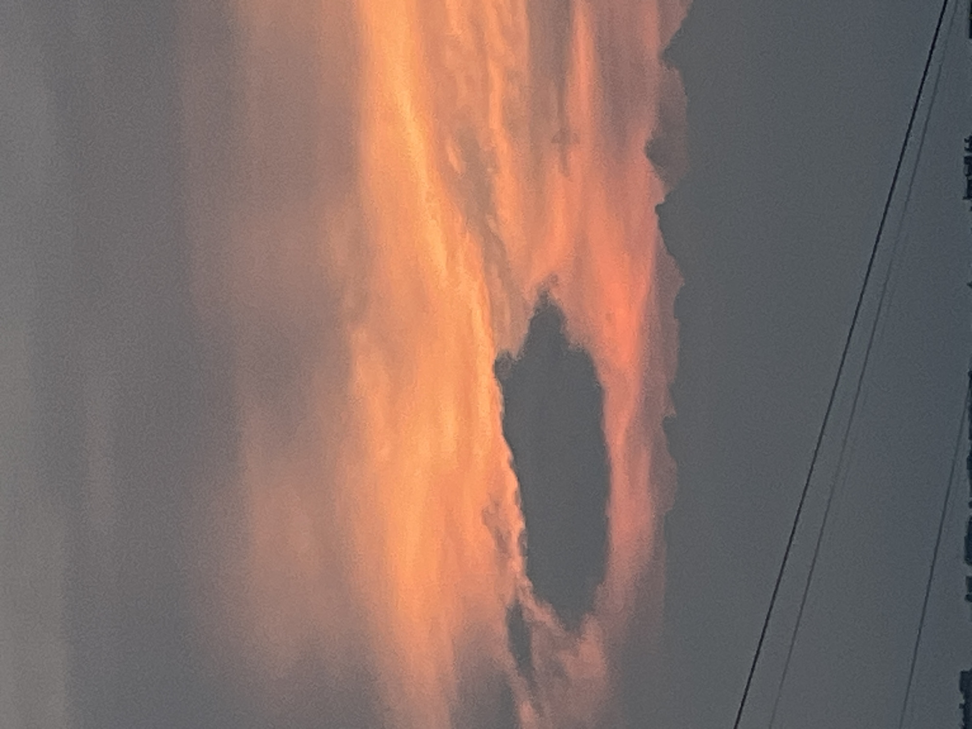 This image is a vertical, medium shot of a sunset over an urban landscape. The sky is filled with a dramatic gradient of colors, transitioning from a deep gray at the top to a vibrant orange and pink towards the horizon. Several clouds are visible, with one prominent, dark cloud formation in the center of the frame appearing to be silhouetted against the brighter sunset hues. In the foreground, there are power lines running horizontally across the lower portion of the image, with a blurry indication of buildings and structures in the far distance, suggesting a city or town setting. The image appears to have been taken at dusk or dawn.
