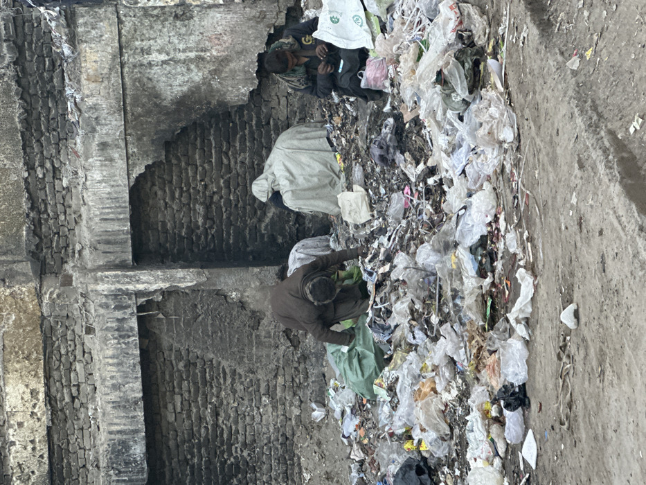 This image depicts an outdoor scene in Quetta City Tehsil, Pakistan, characterized by a substantial accumulation of refuse against a backdrop of urban infrastructure. The background features a severely degraded brick wall structure with crumbling mortar, exposed brickwork, and sections of dark, stained concrete or plaster, including a recessed archway or opening. The ground in the immediate foreground is an unpaved, uneven dirt surface.
A large mound of mixed refuse, predominantly consisting of clear and colored plastic bags, discarded packaging, and various household waste items, occupies the central and lower portions of the frame. Three individuals are present within this refuse.
On the left, an adult male, dressed in dark brown traditional attire, is crouched forward, actively sifting through the trash mound with his hands. A green plastic bag is visible near his position.
In the center, a figure is seated or squatting, entirely enveloped in a light-colored, possibly off-white, sheet or blanket. This individual's actions are not discernible due to the covering.
On the right, another adult male, wearing dark clothing and a multi-colored scarf, is seated amongst the refuse. He is observed manipulating small items with both hands. A white plastic bag with green lettering and a smaller pink plastic bag are situated next to him. The overall scene suggests scavenging for materials within a neglected urban environment.