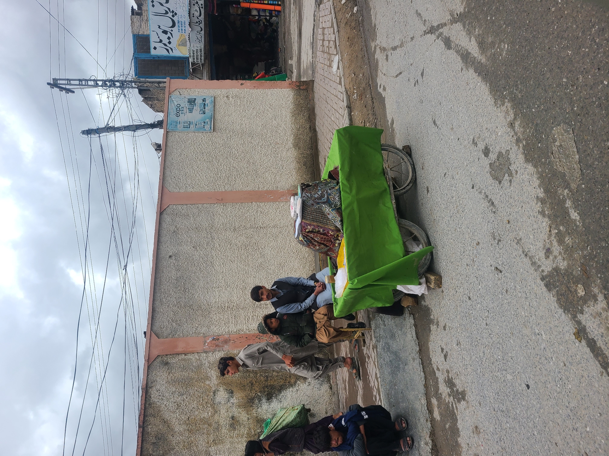 The scene shows a group of people gathered on a sidewalk in Quetta City Tehsil, Pakistan. A pushcart covered with a green tarp stands beside a wall, displaying fabrics. Several men are either sitting, standing, or lying down near the cart. One person is sitting on a small stool. The area appears to be a commercial district, with a storefront visible in the background. Overhead are numerous power lines against a cloudy sky. The street is paved, and there are water stains on the sidewalk.