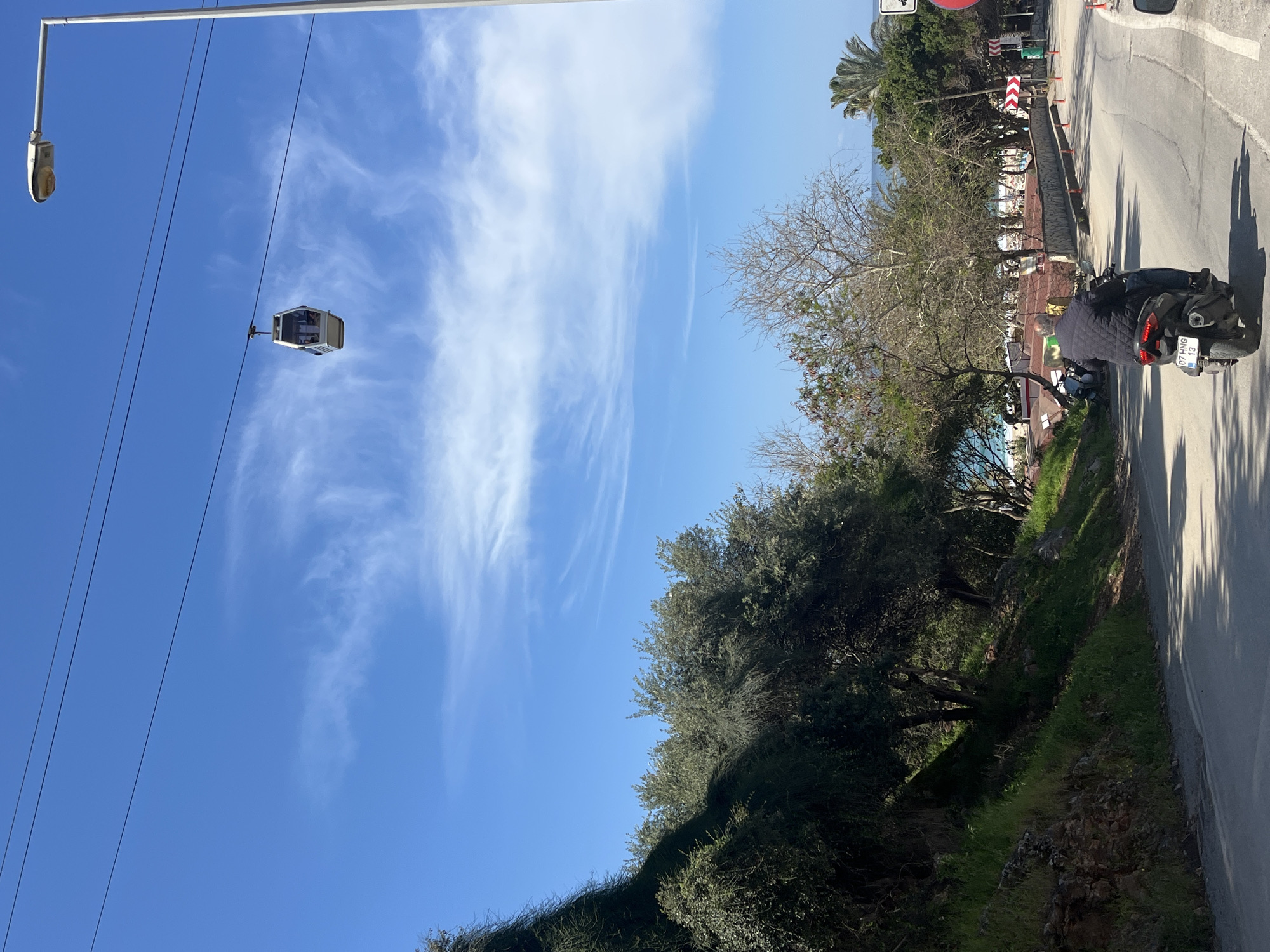 The image captures a street scene in Alanya, Turkey, featuring a cable car cabin suspended in the air on a clear day with wispy clouds. The foreground includes dense vegetation lining the side of the road, with trees and shrubs obscuring some of the view. A motorcycle is parked on the side of the road, and a person is standing nearby. In the background, buildings and the sea can be glimpsed through the trees. The road is asphalt with a curb. A street light is visible to the upper left. The license plate on the motorcycle reads "07 HNG 13".
