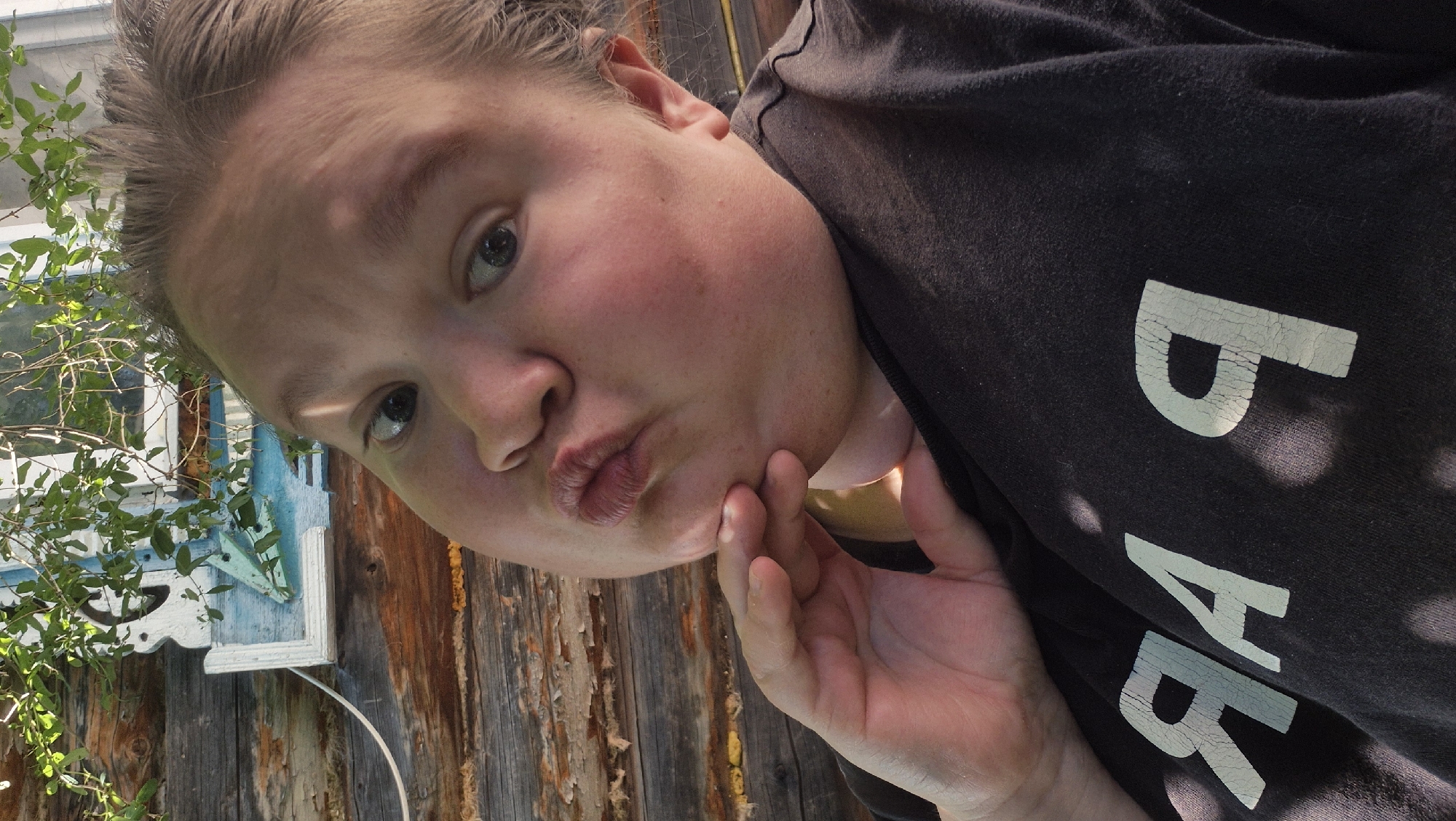 This is a close-up, slightly tilted selfie of a person with light-colored hair styled upwards, taken outdoors during the daytime. The person's face is the primary subject, with their gaze directed towards the camera. Their cheeks are flushed, and their lips are puckered in a kissing pose. One hand is resting on their chin, and the other is obscured.
The background consists of a weathered wooden wall, suggesting a rustic or rural setting. To the left of the frame, a portion of a white and blue window frame is visible, adorned with decorative carvings and surrounded by lush green foliage. This combination of elements hints at a rural Russian dacha or house in the village of Doynaya, Russia.
The person is wearing a dark t-shirt with light-colored text printed on it. The visible letters suggest the word "BABY," with parts of "B" and "A" clearly legible. The text appears cracked and faded, indicating wear on the garment. The sunlight creates strong highlights on the person's face and the t-shirt, suggesting a bright, sunny day. The overall mood appears casual and playful, typical of a personal selfie.