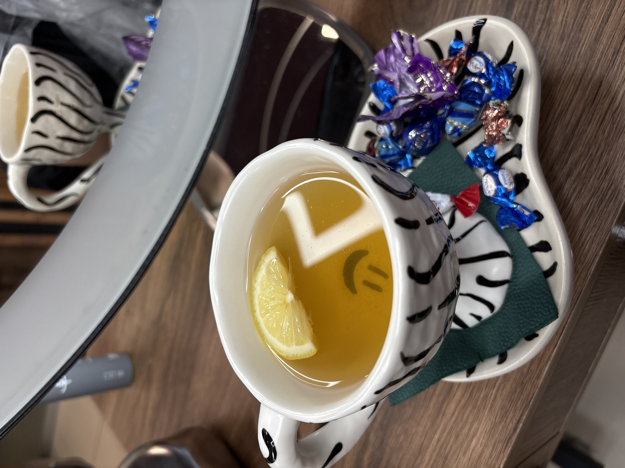 A close-up shot shows a white mug with black zebra-like stripes containing a light brown liquid and a lemon slice. The mug is placed on a saucer that also has black stripes and holds several foil-wrapped candies in blue, red, and purple. A green napkin is tucked under the mug. The scene is set on a wooden surface, and a reflection in the background shows a blurred interior with striped patterns. No specific location in Birobidzhan, Russia, is discernible from the image. There are no people or actions depicted.