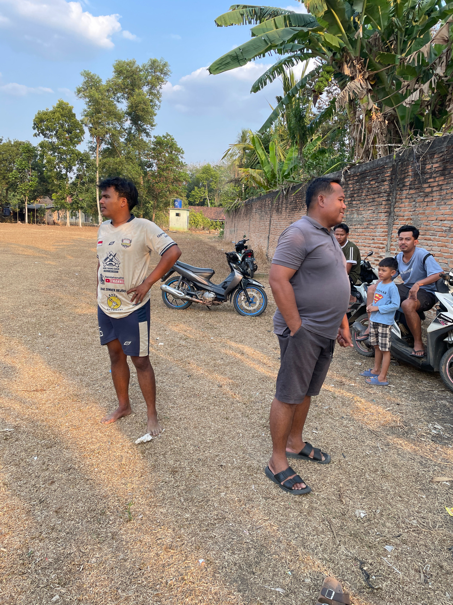The image depicts an outdoor scene in Kagungan Ratu, Indonesia, characterized by dry, straw-like ground cover and a brick wall bordering the right side of the frame. The sky is clear with scattered clouds, and the lighting suggests it is late afternoon or early evening.
In the foreground, a man wearing a light-colored t-shirt with various logos and dark shorts stands facing left, with his right hand on his hip. His feet are bare. To his right, a larger man in a grey polo shirt and dark shorts stands with his hands in his pockets, facing right. Behind him and to the right, two other men and a young boy are gathered. One man wears a green and white striped long-sleeved shirt, another wears a blue t-shirt, and the boy is dressed in a light blue long-sleeved shirt with a Superman logo and plaid shorts.
Several motorcycles are present in the background. One is a dark-colored scooter parked behind the man in the t-shirt and shorts. Another is a white scooter to the right, partially obscured by the group of people. In the far background, trees and some simple structures are visible. Banana trees with large leaves are prominent near the brick wall. The overall atmosphere appears casual, with the individuals seemingly engaged in conversation or observing something.