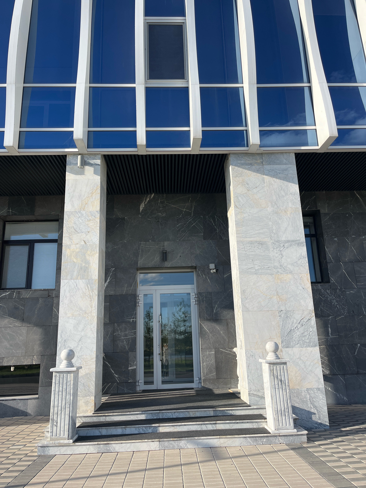 The image shows the exterior of a modern building with a facade of dark gray stone and large blue windows. The entrance is framed by two tall, white marble columns. A glass door leads into the building. The ground in front of the entrance is paved with light-colored tiles. Two white decorative pillars with spherical tops stand on either side of the entrance steps. The building appears to be located in a city, with the sky visible above. The architecture suggests a contemporary commercial or public building. The specific location in Astana, Kazakhstan, is not visually determinable from the image alone.