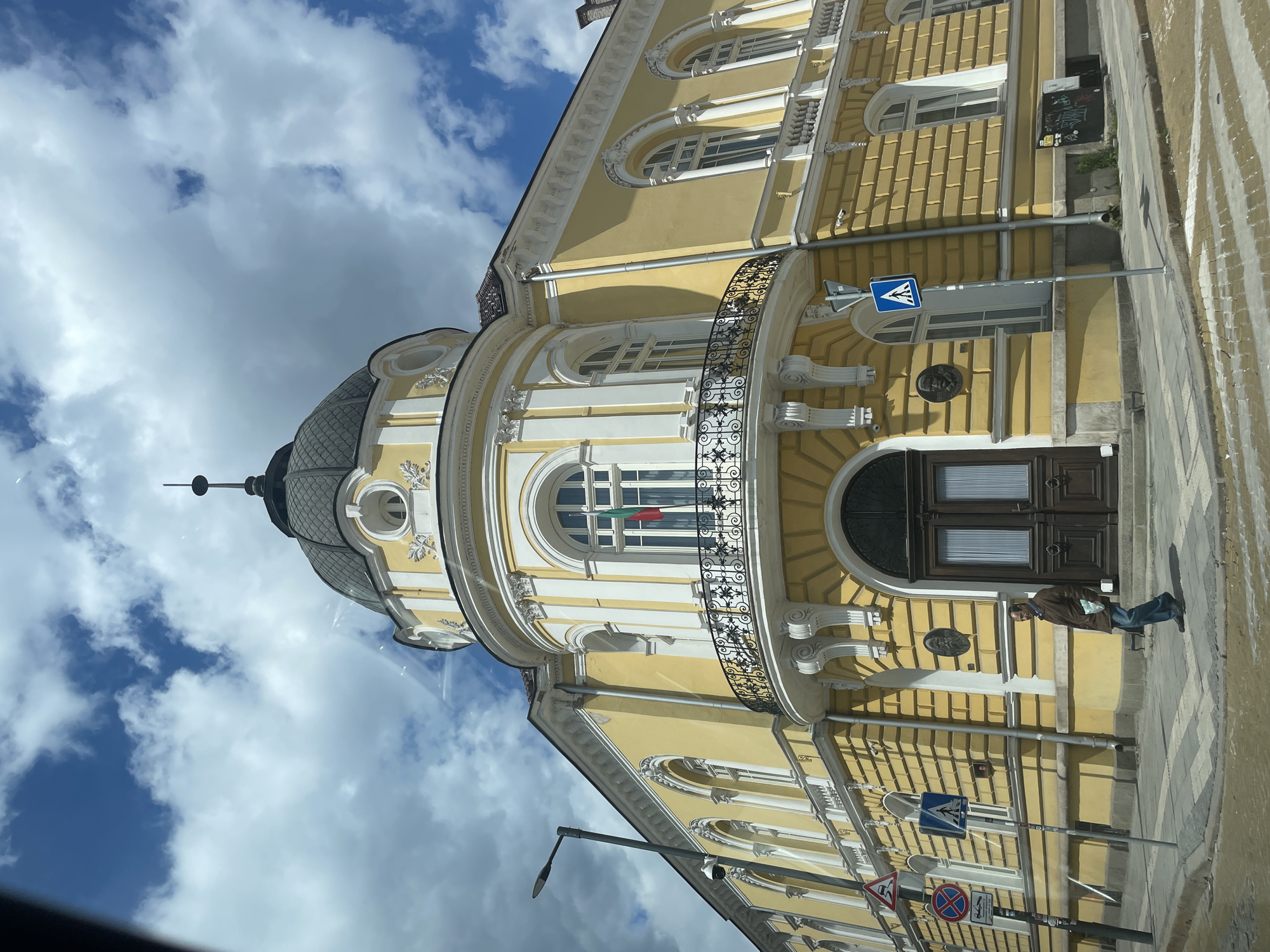 Here is a detailed description of the image:
The image shows the facade of a yellow building with white architectural details in Sofia, Bulgaria. The building is ornate, featuring arched windows, a balcony with wrought iron railings, and a domed cupola. A Bulgarian flag is visible hanging in one of the arched windows.
The street level includes a pedestrian crossing sign, other road signs, and a street light.
A person is standing on the sidewalk near the building entrance. The sky is visible in the background and is partly cloudy.