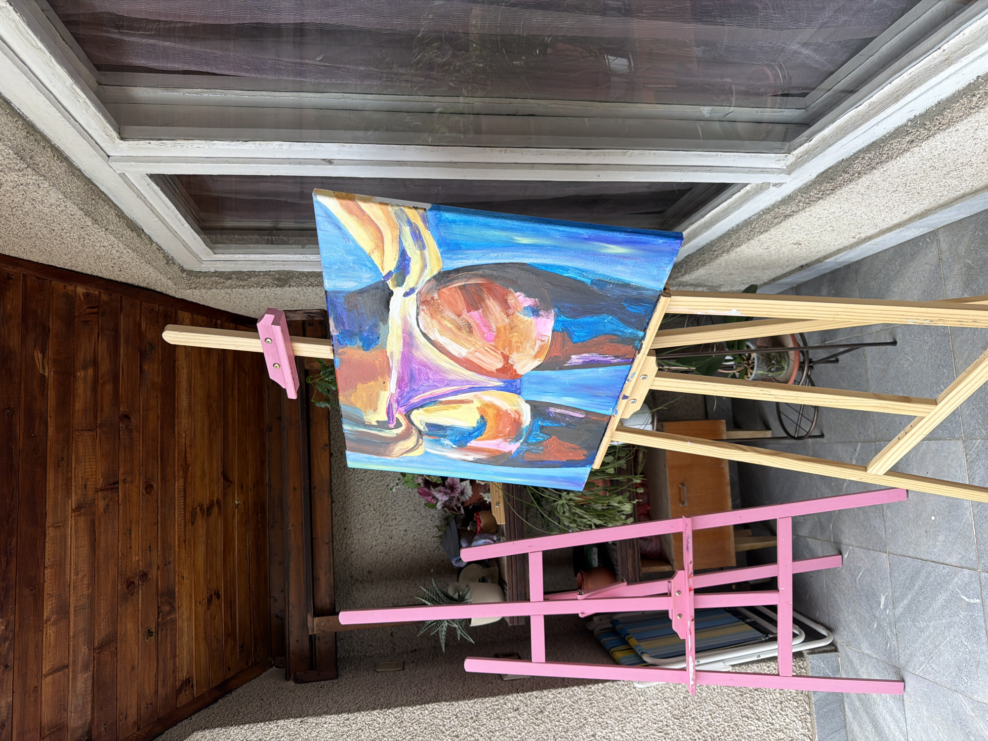 A painting of buttocks, rendered in abstract, bold strokes of color, is displayed on a wooden easel on a tiled patio. The painting features vibrant blues, purples, pinks, and oranges, with thick impasto techniques evident. The background of the scene reveals a textured, light-colored wall, a wooden ceiling, and a window with sheer purple curtains. To the left of the main easel, a bright pink easel stands, holding nothing, and a stack of striped beach chairs are visible behind it. Several potted plants are arranged on shelves and a small table in the background, adding a touch of greenery to the outdoor setting. The overall impression is one of an artist's workspace or an outdoor exhibition, possibly located in a residential area in Svilengrad, Bulgaria, given the architectural style.