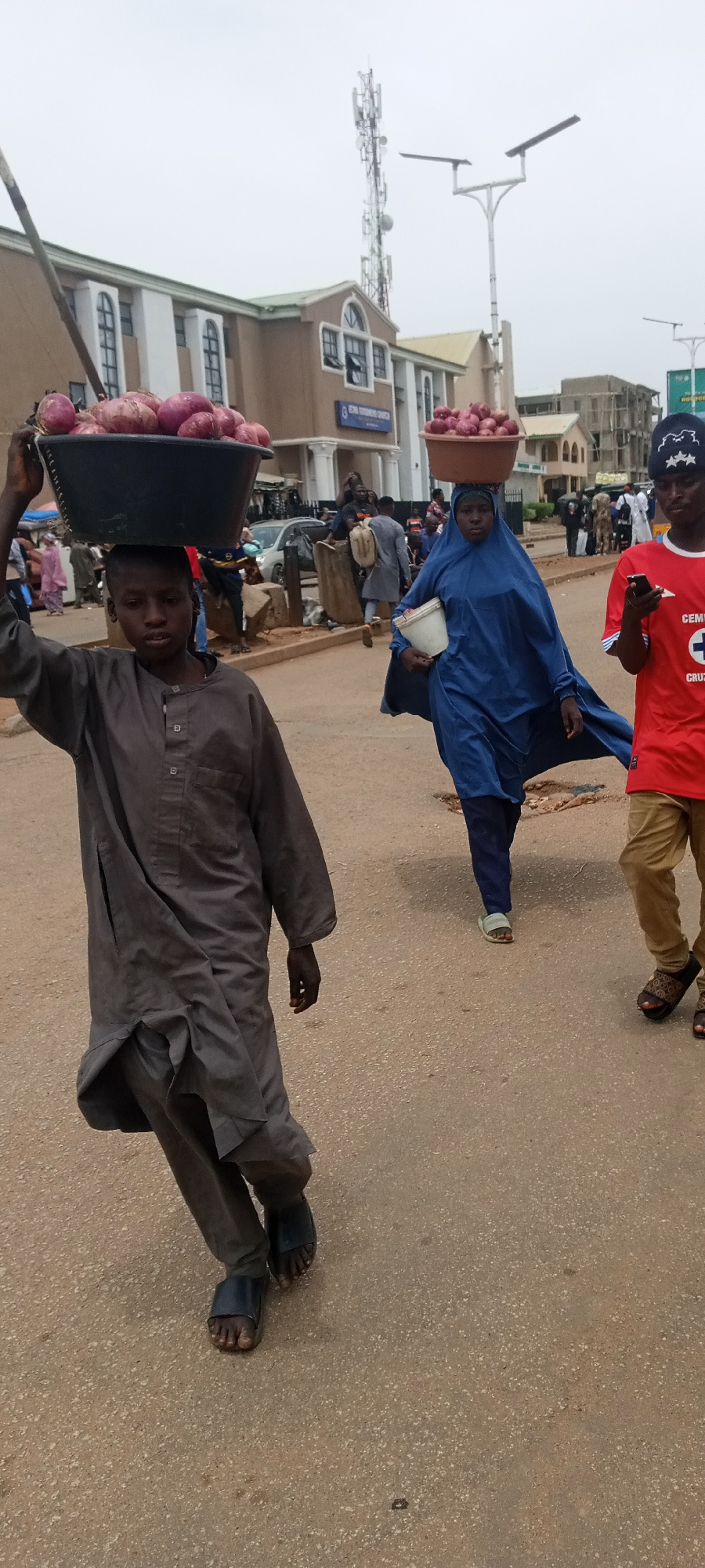 In Jos, Nigeria, a young person wearing a full-length gray gown and dark sandals carries a black tub filled with red onions on their head, facing forward. To their right, a woman in a full-length blue garment and head covering carries a white container, also with a tub of red onions balanced on her head. A third person in a red t-shirt and khaki pants walks to the woman's right, looking at a mobile phone. The scene takes place on a paved street; other individuals and vehicles are in the background. A building with white arched windows and a sign indicating "ECWA GOODNEWS CHURCH" is visible behind the first person. A communication tower and street lights are in the background as well.