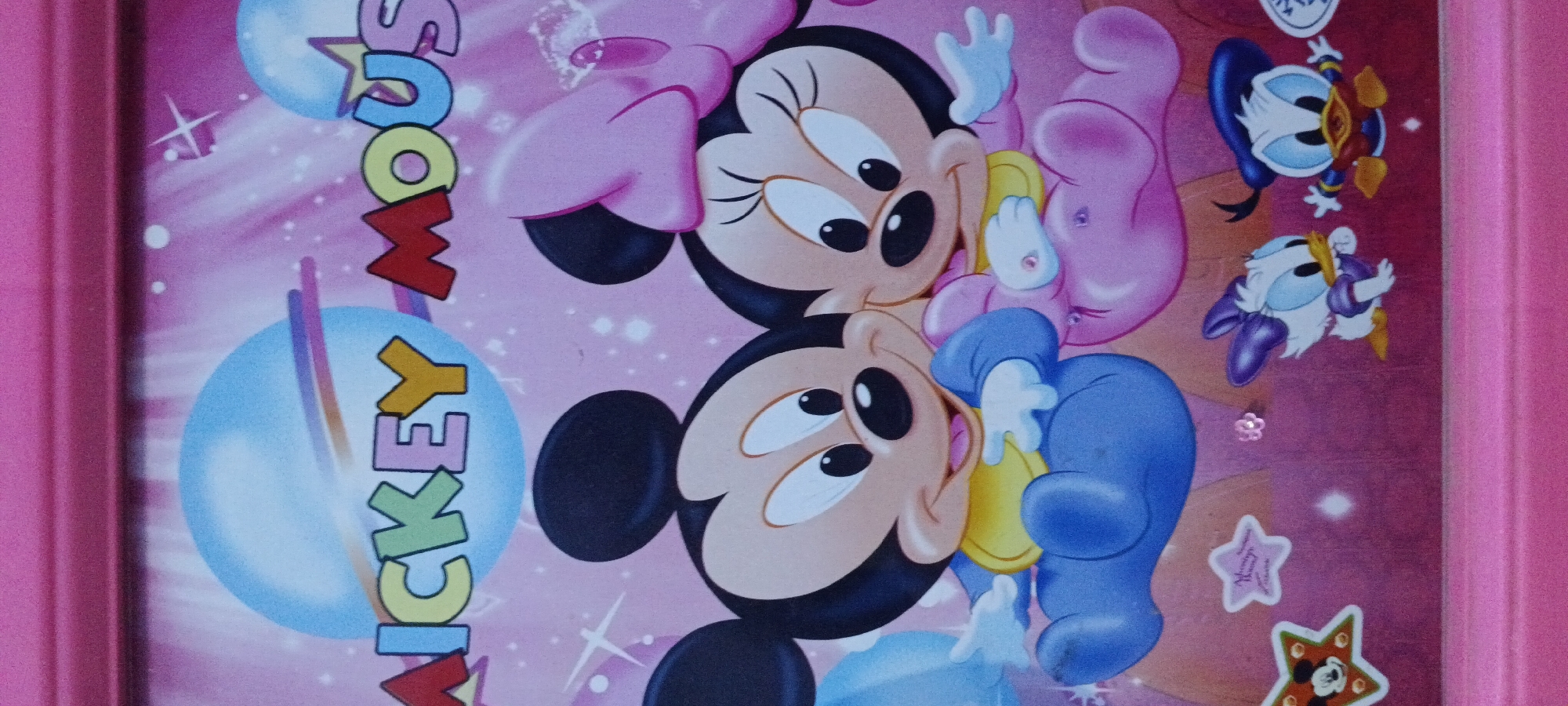 The image is a close-up of a pink background featuring cartoon characters from Disney's Mickey Mouse franchise. In the center, Mickey Mouse and Minnie Mouse are depicted as babies, with Mickey wearing a blue onesie and a yellow bib, and Minnie in a pink outfit with a pink bow. Both have large heads with black ears and big eyes. To the bottom left, there is a star-shaped sticker with Mickey Mouse's face. To the right of the central characters, Daisy Duck and Donald Duck are also shown as babies. Daisy is wearing a purple dress and Donald is in a blue outfit with a blue hat. The background has a pink gradient with light blue spheres and sparkling star shapes. The image appears to be a print on a toy or decorative item, as indicated by the solid pink border at the top and bottom. There is no visible information about the location being Rejing, Indonesia.