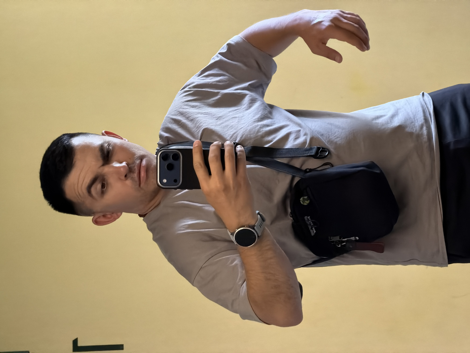 A medium shot captures a man taking a mirror selfie in what appears to be a room with a pale yellow wall. The man has short dark hair and is looking at his phone, which he holds with his right hand. He is wearing a light gray t-shirt, a black crossbody bag, and black pants. A dark-colored watch with a round face is visible on his left wrist. His left arm is bent at the elbow, with his hand in a relaxed, open position. The background shows a faint dark green 'T' shaped object on the left side of the wall. The scene is indoors and appears to be a private space, possibly a home or a gym, given the presence of what looks like a fitness tracker. There is no specific indication of the city of Izhevsk, Russia.