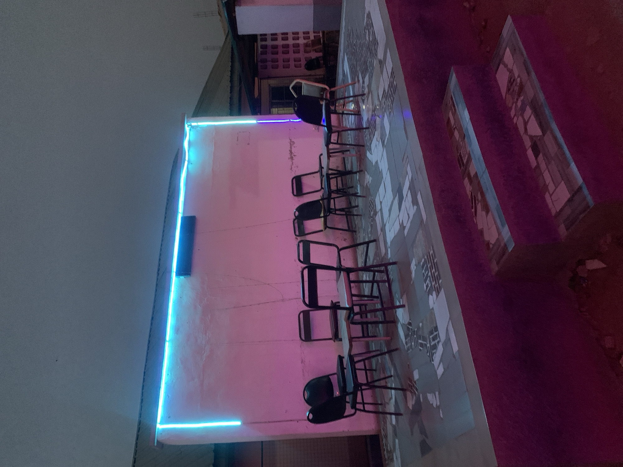The image shows a nighttime scene on a rooftop in Kabong, Nigeria. A row of about ten black metal chairs is lined up against a wall that is illuminated with pink light and blue neon. The chairs are on a tiled surface. A building is in the background. The sky is overcast.