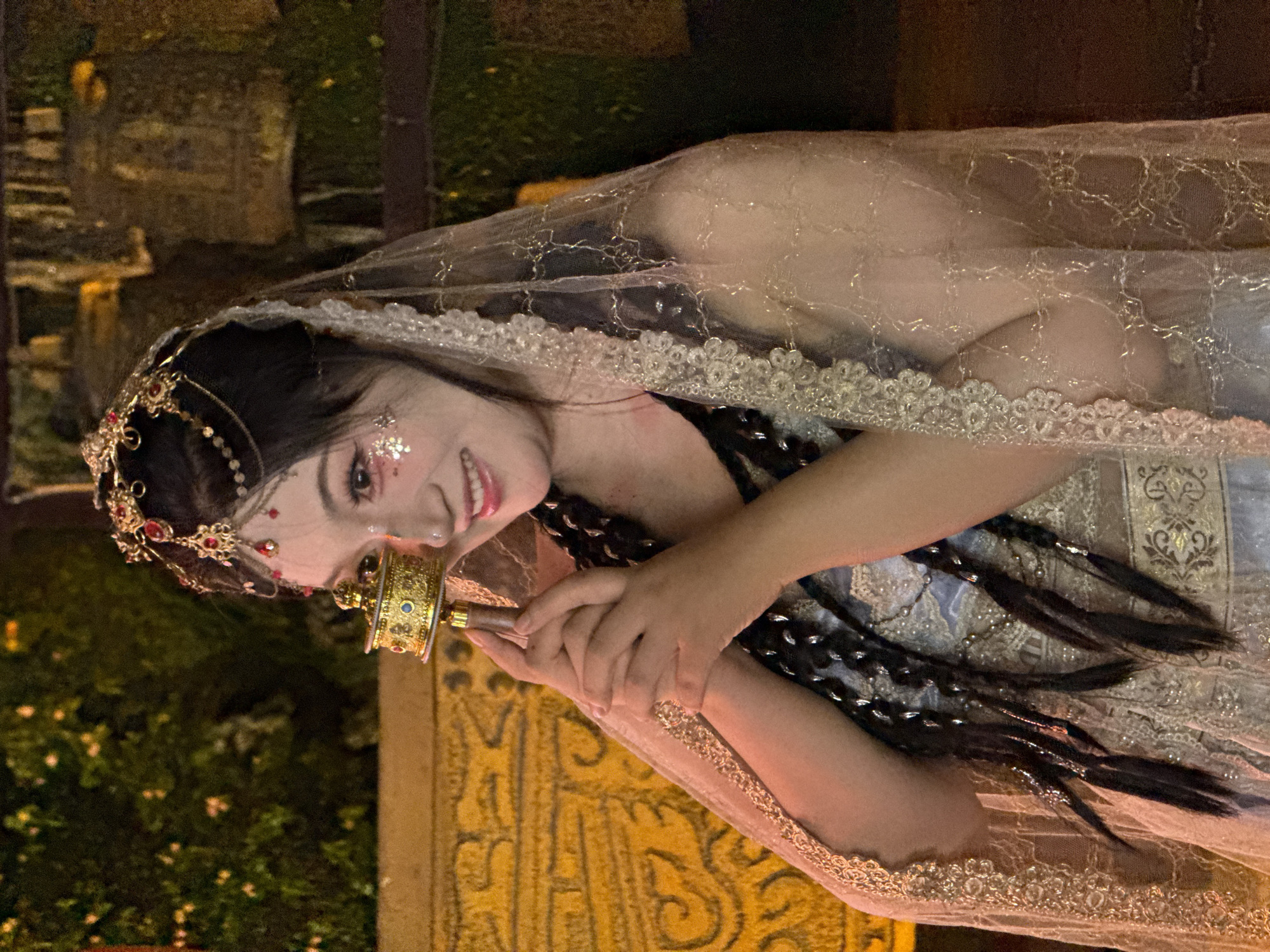 A woman is holding a hand drum outdoors. The woman is wearing a sheer veil with lace trim, a headpiece with jewels, and has glitter on her face. She has long, dark hair styled in braids. She is holding a decorated hand drum in both hands. The background shows out-of-focus vegetation and building details. The location appears to be the Yuzhong District in China.