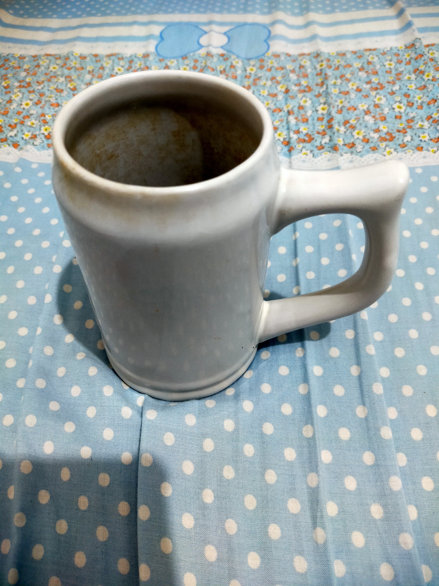 A white ceramic mug with a light brown interior sits on a light blue tablecloth with white polka dots. The mug is positioned at an angle, with its handle facing to the right. Behind the mug, the tablecloth transitions to a section with a floral pattern featuring small white and orange flowers on a blue background, bordered by a white lace-like trim. Further back, there's a section of horizontal blue and white stripes, with a light blue cloud shape visible on the leftmost stripe. The lighting suggests an indoor setting. The mug is the primary subject, and its placement on the patterned tablecloth creates a simple still life composition. No people or actions are depicted.