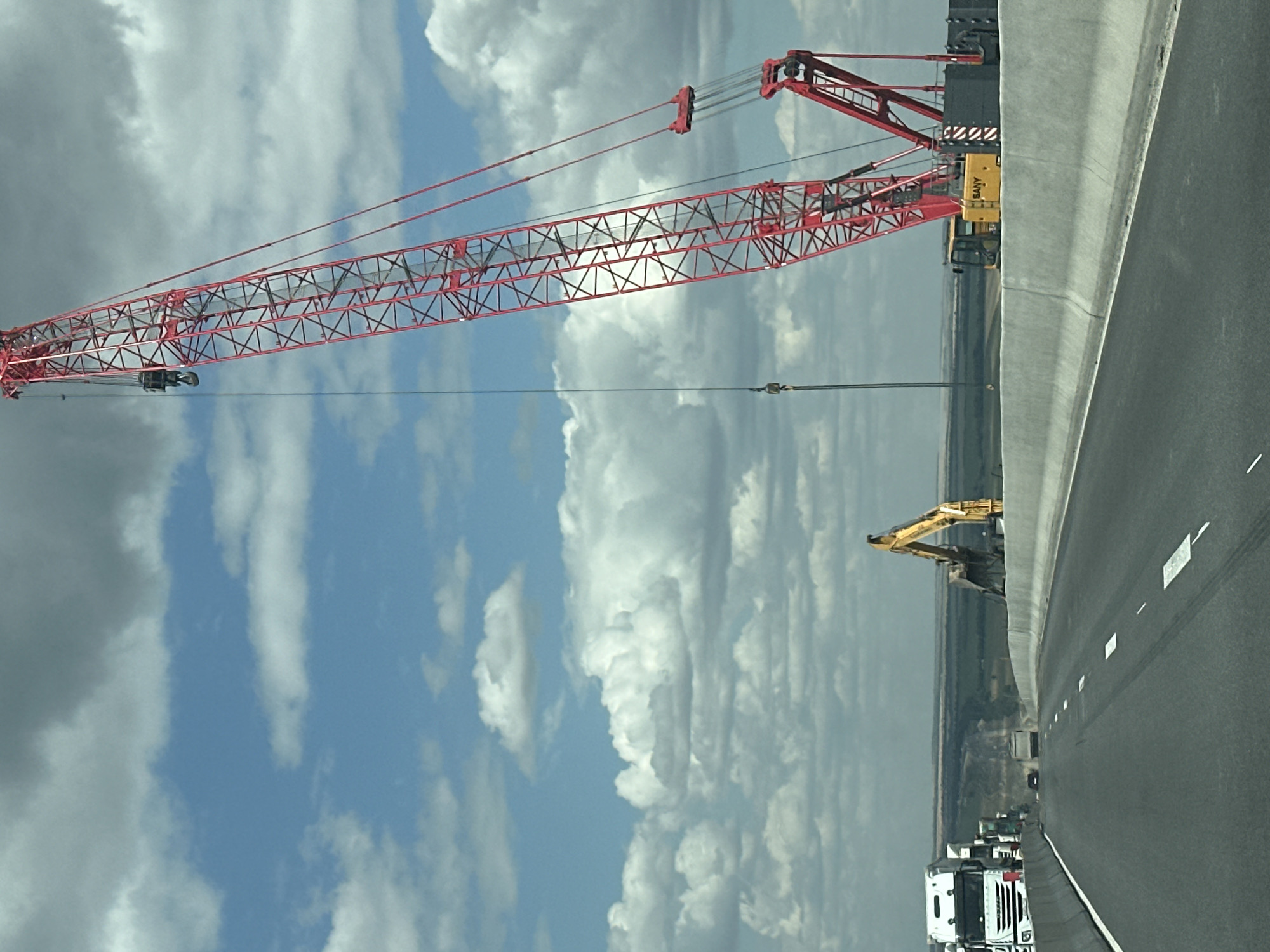 This image shows a construction site on a highway, likely a bridge under construction. A large red crane dominates the right side of the frame, with its boom extended upwards and to the right. A yellow excavator is visible in the middle ground, positioned behind a concrete barrier. Several white trucks are parked on the left side of the highway, suggesting ongoing work. The scene is set against a backdrop of a cloudy sky and a distant, flat landscape. The highway itself appears to be a multi-lane road with clear lane markings. The presence of construction equipment and barriers indicates that this is an active work zone. The specific location in Donleskhoz, Russia is not visually evident from the image itself, but the context suggests it is a remote or developing area. There are no people visible in the image.
