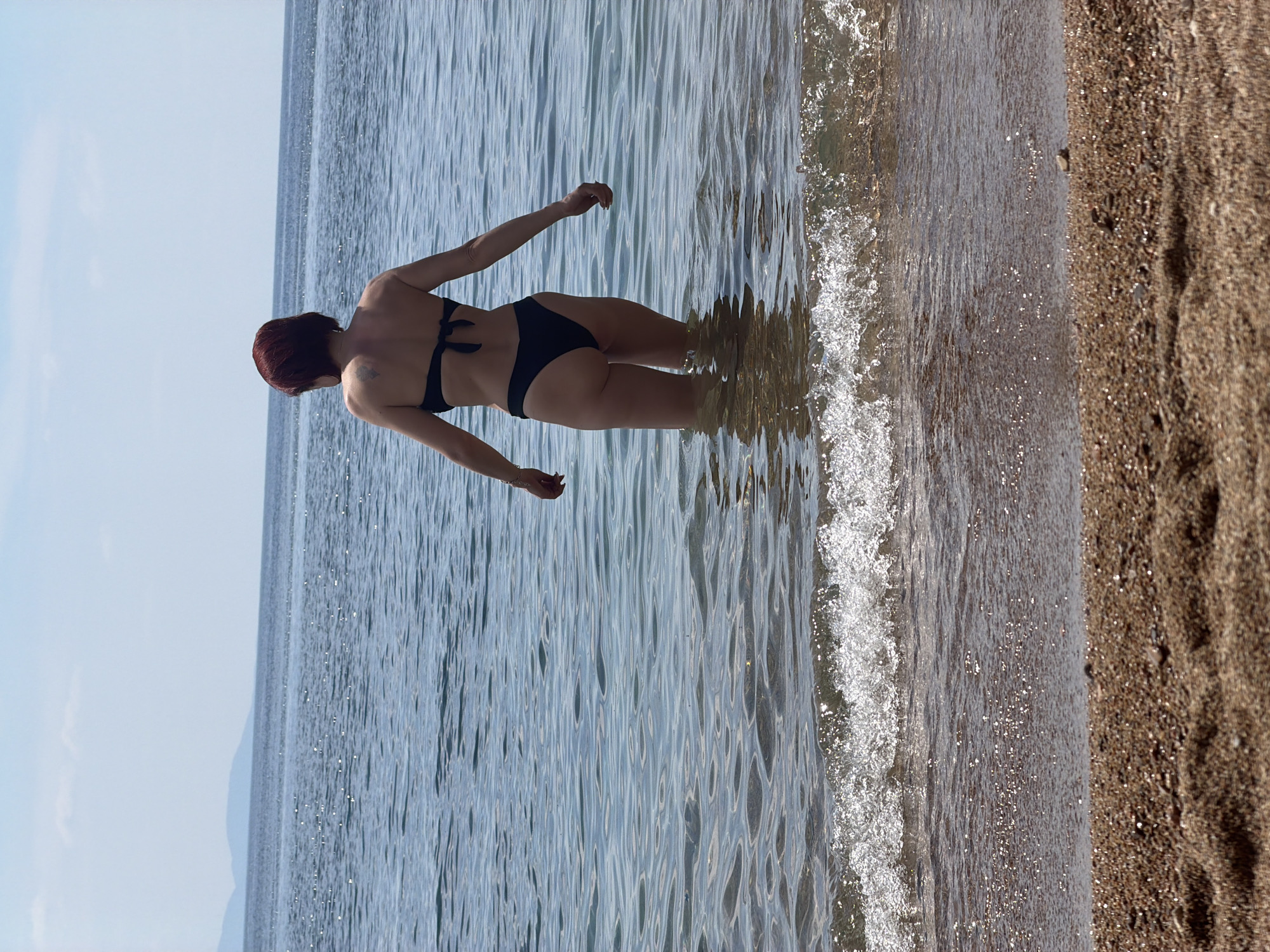 The image shows a woman standing in the sea. She is facing away from the camera. The woman has red hair and is wearing a black bikini. The sea is calm and clear. The beach is made of sand. The image appears to be taken in Budva, Montenegro, given the location mentioned.