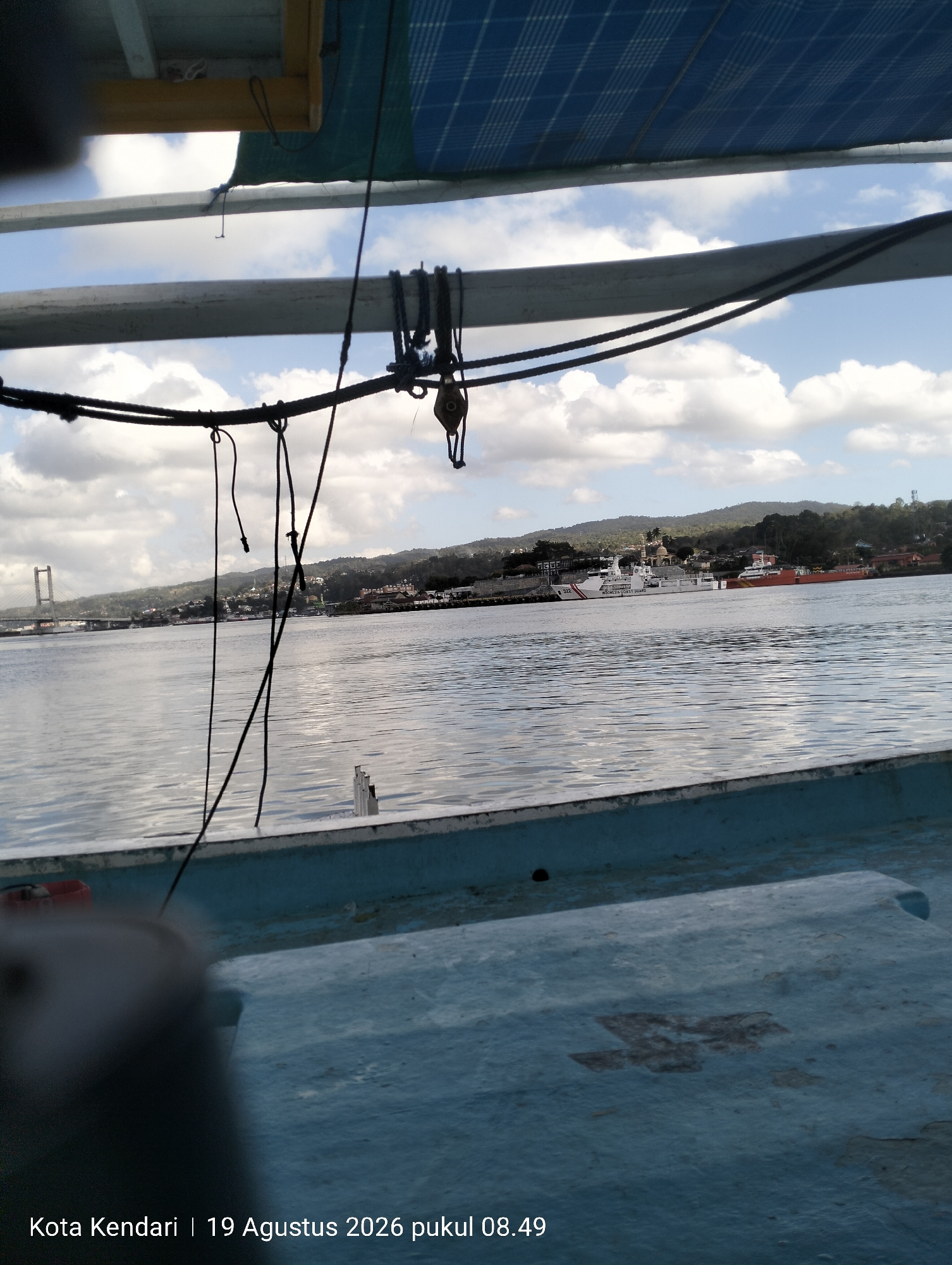The image shows a view from a boat on the water, looking towards a city on the coast. In the distance, a suspension bridge is visible. Several ships are docked near the shore, including what appears to be a coast guard vessel. The sky is partly cloudy, with a blue sky visible above the clouds. The foreground of the image shows part of the boat the photo was taken from, with some ropes and a pulley system. The bottom of the image contains a timestamp and location: "Kota Kendari | 19 Agustus 2026 pukul 08.49". This indicates the location is Kendari, Indonesia, on August 19, 2026, at 8:49 AM.