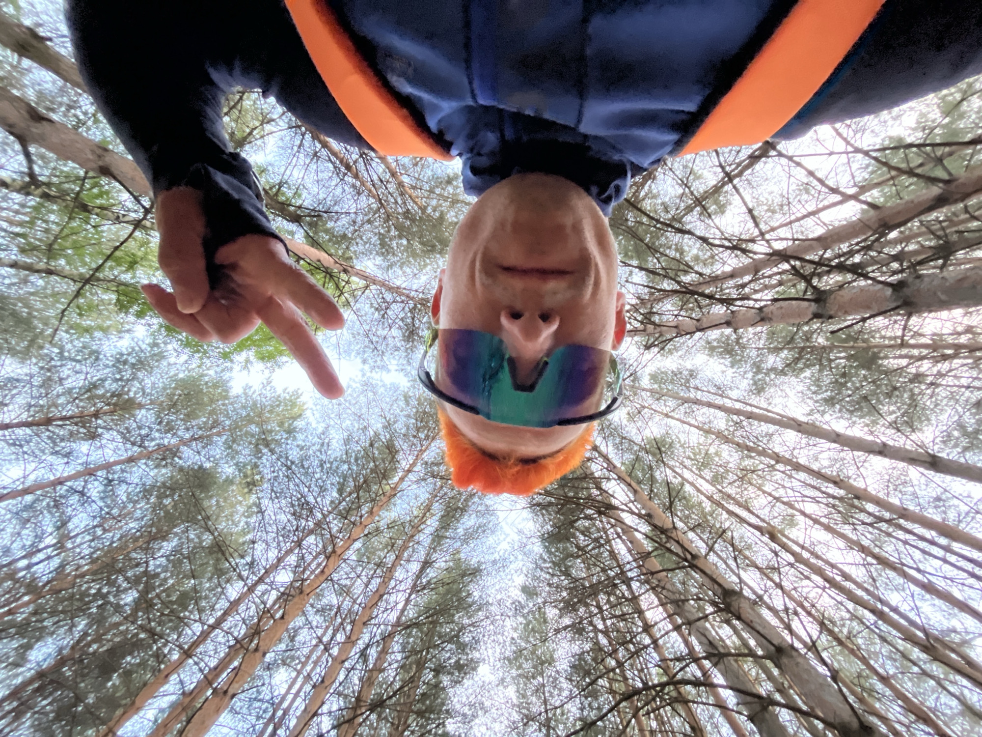 A low-angle selfie photograph shows a person with orange hair and iridescent sunglasses, oriented upside-down. They are making a peace sign with their right hand. They are wearing a blue shirt with orange straps. The background consists of tall trees angled towards the center of the frame, suggesting a forest setting. The location is in the city of Chaikovsky, Russia.
