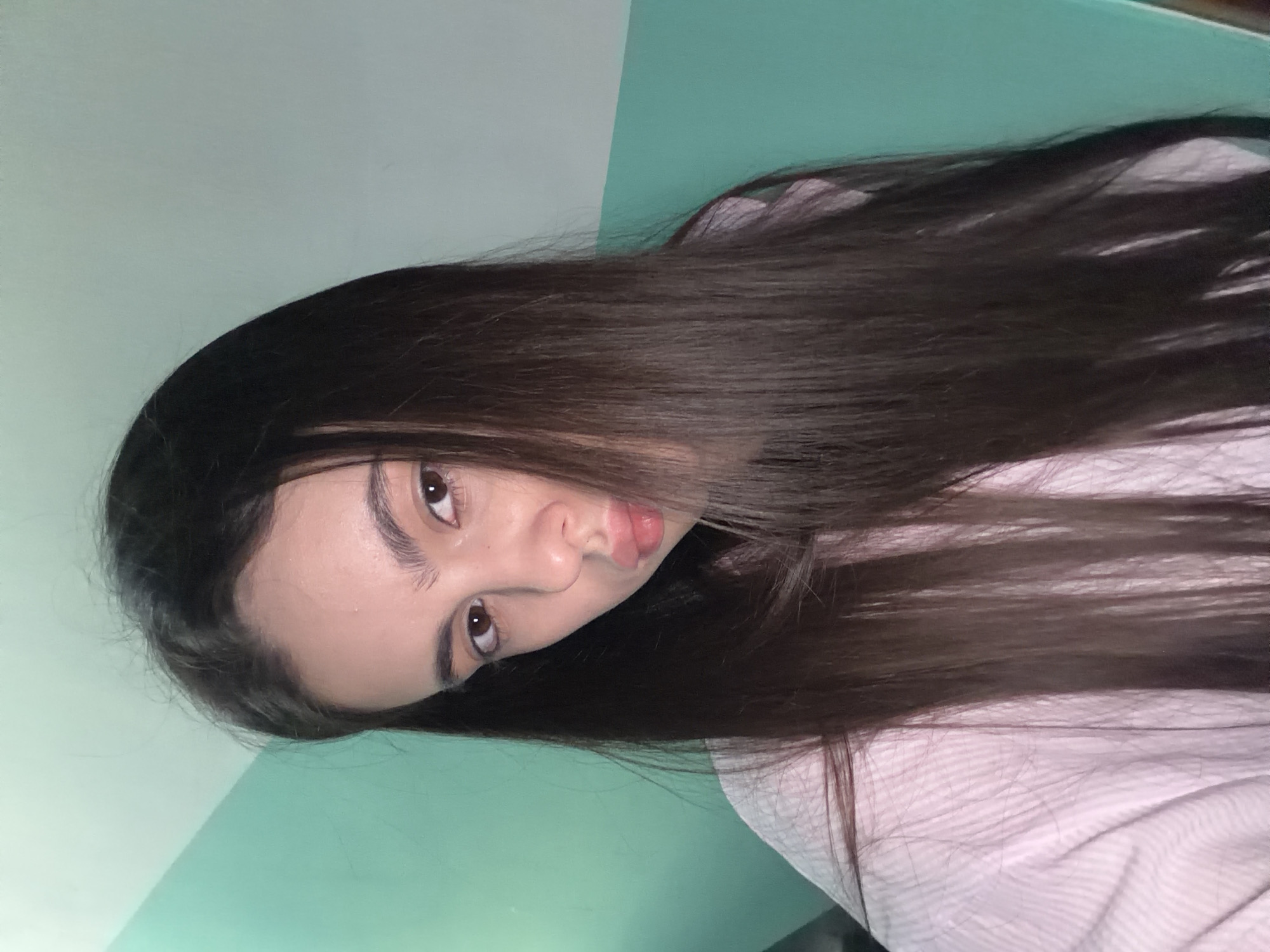 A rotated indoor selfie shows a young woman with dark brown hair against a backdrop of light blue and white walls. She wears a pink patterned shirt or pajama top. Her face is angled towards the camera with a neutral expression and slightly pursed lips. Her eyes are brown. There is no verifiable information connecting the photo to the city of Chaikovsky, Russia.