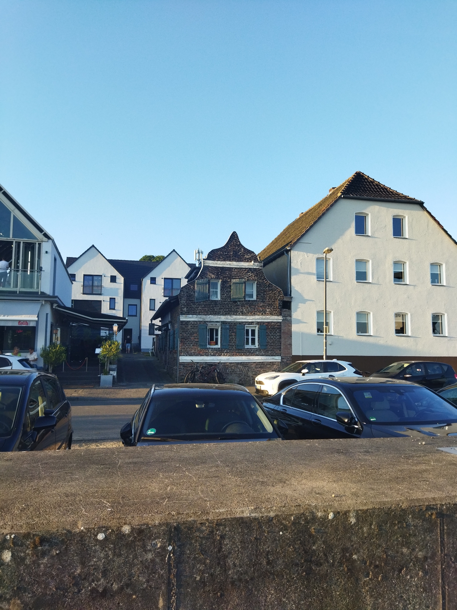The image depicts a street scene in Leverkusen, Germany, bathed in the warm light of late afternoon sun under a clear blue sky. In the foreground, a textured concrete barrier runs horizontally across the frame, partially obscuring several parked cars. Two dark-colored sedans are visible, one in the lower left and another in the lower center, their roofs and hoods peeking over the barrier.
Beyond the barrier, the street is lined with buildings of varying architectural styles. To the left, a modern building with a glass facade and a balcony overlooks the street. Adjacent to it is a restaurant with outdoor seating covered by an awning. Further down the street, a row of residential buildings stands, featuring white exteriors and dark tiled roofs. A distinct, older, narrow brick building with a stepped gable roof and green shutters is nestled between the modern structures, adding historical character to the scene.
Several cars are parked along the street, including a white crossover SUV and a black hatchback. A tall street lamp stands near the white residential building. A person in dark clothing can be seen walking away from the camera on the street. In the background, trees are visible, suggesting a green urban environment. The overall impression is one of a calm, well-maintained urban area.