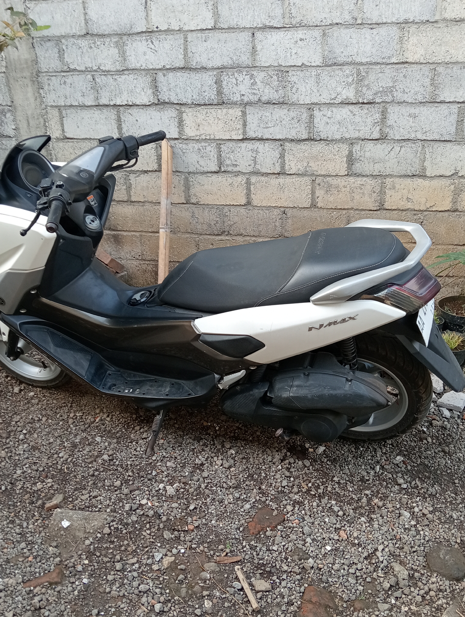 A white and black Yamaha NMAX scooter is parked on a gravel surface against a rough, light-colored brick wall. A wooden stake is propped against the scooter's handlebars. The scooter is viewed from the side, showing its seat, rear wheel, and engine casing. The environment appears to be an outdoor area with some foliage and scattered debris. The image was taken in Krajan Kidul, Indonesia.