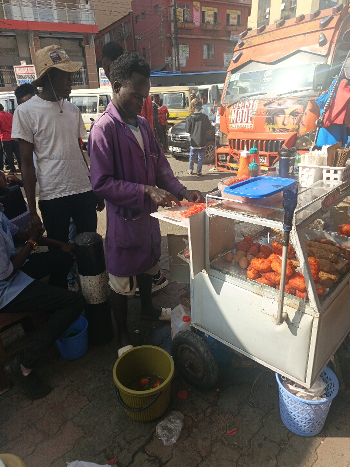 The image captures an outdoor street scene in a busy commercial area of Nairobi, Kenya, characterized by street vending and public transportation. In the foreground, a dark-skinned adult male, wearing a purple long-sleeved jacket over a light shirt and white shorts, stands facing right. He is actively chopping red food items, likely tomatoes, on a small white cutting board affixed to a mobile food cart. His hands are dark and engaged with a knife.
To his immediate left, a second dark-skinned adult male stands, observing the food preparation. He wears a white t-shirt, dark trousers, and a light brown wide-brimmed bucket hat, with a dark cord around his neck. In the bottom-left, a third dark-skinned adult male is partially visible, seated on a low stool, wearing a light blue collared shirt and dark trousers. A blue plastic bucket is near his feet.
The white mobile food cart, positioned on the right, features a glass display section containing multiple reddish-orange cooked food items, several raw eggs, and various condiments in plastic bottles on its top surface, alongside white plastic organizers and a blue lidded container. On the ground adjacent to the cart, a green plastic bucket holds water and two red tomatoes or similar produce. A light blue plastic basket and scattered food scraps are also present on the brick-paved ground.
The background reveals a bustling street with several dark-skinned pedestrians and multiple vehicles. A prominent orange public service vehicle (matatu) is visible, heavily customized with text including "BEAST MODE ACTUATED" and a graphic of a woman's face on its front. Other minibuses and a black sedan are also present. Multi-story buildings, including a reddish-brown structure, line the background under direct sunlight, casting distinct shadows.