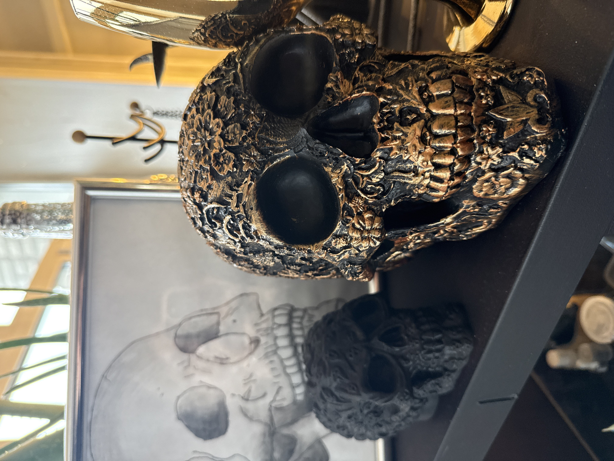 A close-up, canted shot depicts a decorative skull and a framed drawing of a skull on a dark shelf. The decorative skull is black and gold, with an ornate design. The framed drawing behind it is black and white. The shelf appears to be made of metal and is black. Other blurred objects are visible in the background. The location in the city of Krasnoyarsk, Russia, cannot be determined from this image.