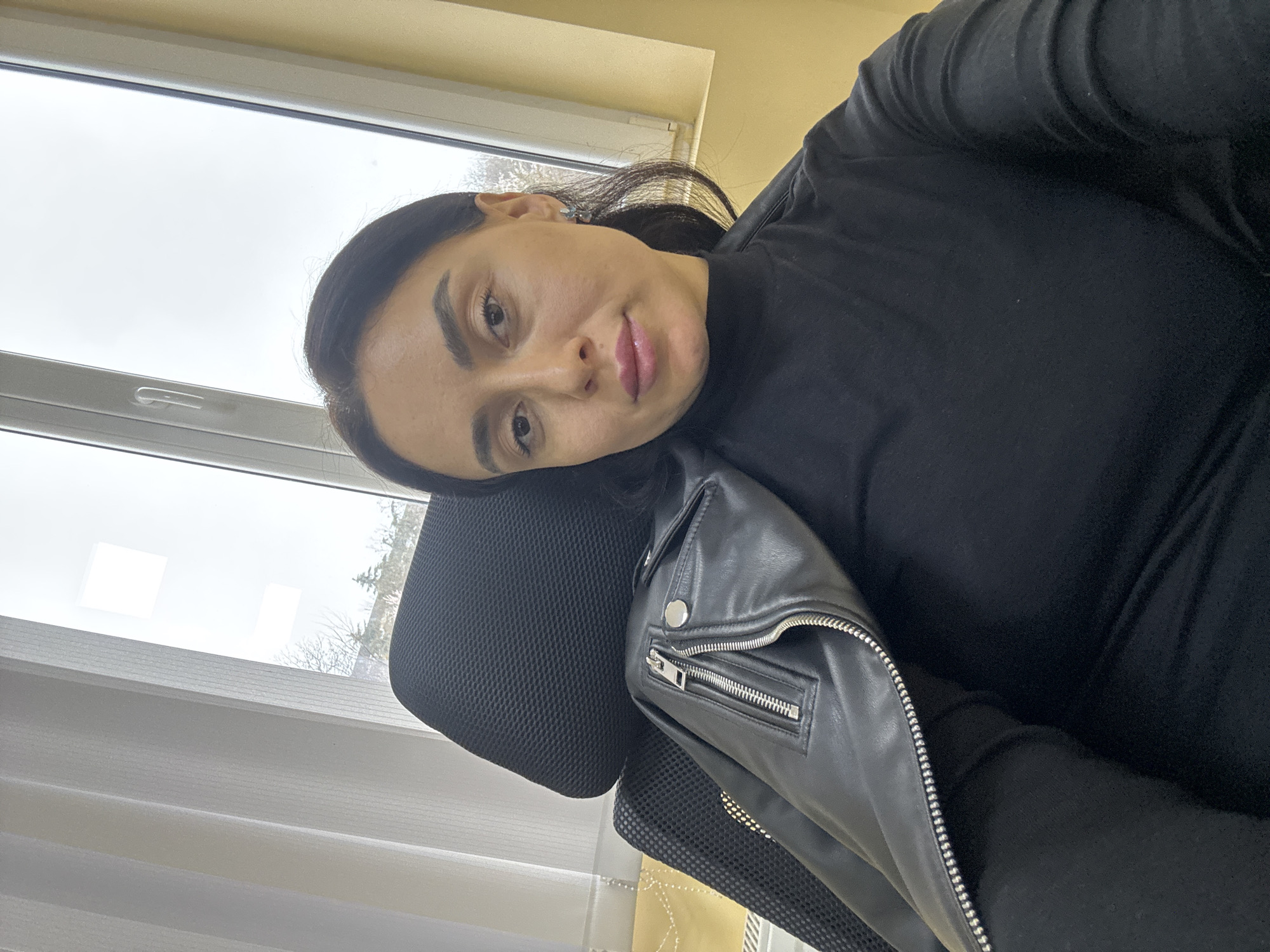 A woman is depicted in a mid-shot selfie, seated in a black mesh office chair indoors. The location is specified as Tbilisi, Georgia. She has dark hair, a fair complexion, and wears a black long-sleeved turtleneck. A black leather or faux leather jacket, featuring silver zippers and snaps, is draped over her left shoulder and the chair back. Small, light-colored earrings are visible. She gazes directly at the camera with a neutral expression. Behind her is a white-framed window, partially covered on the left by vertical white blinds. Through the window, the outdoor environment shows a bare tree against an overcast sky, with some green foliage in the distance. The interior wall to the right of the window is a light yellow or beige color. A white radiator is partially visible beneath the window on the far left.