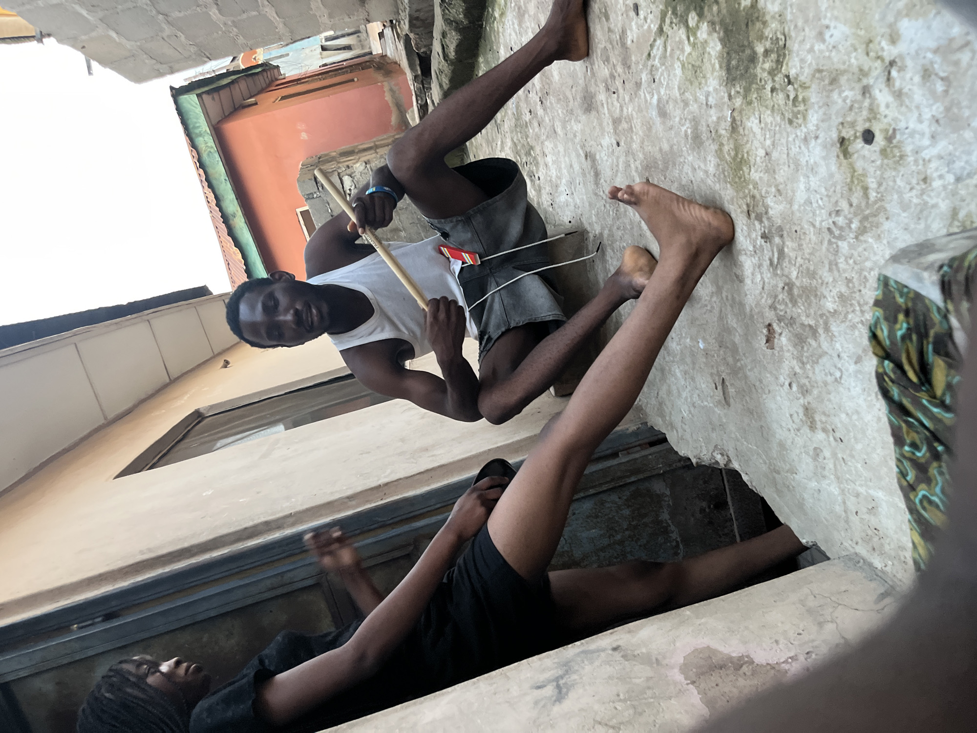 Two individuals are present in an outdoor urban alleyway. On the left, a young woman with braided hair, wearing a black t-shirt and shorts, stands near a metal doorway. Her left leg is extended forward. To her right, a young man sits on the ground, facing the camera. He is wearing a white tank top and grey shorts. He holds a long, thin wooden stick in his hands, with his right hand on the stick and his left hand resting on his lap. The ground is paved and appears worn. Buildings with various colored facades and corrugated metal roofs are visible in the background. The sky is bright and overcast.