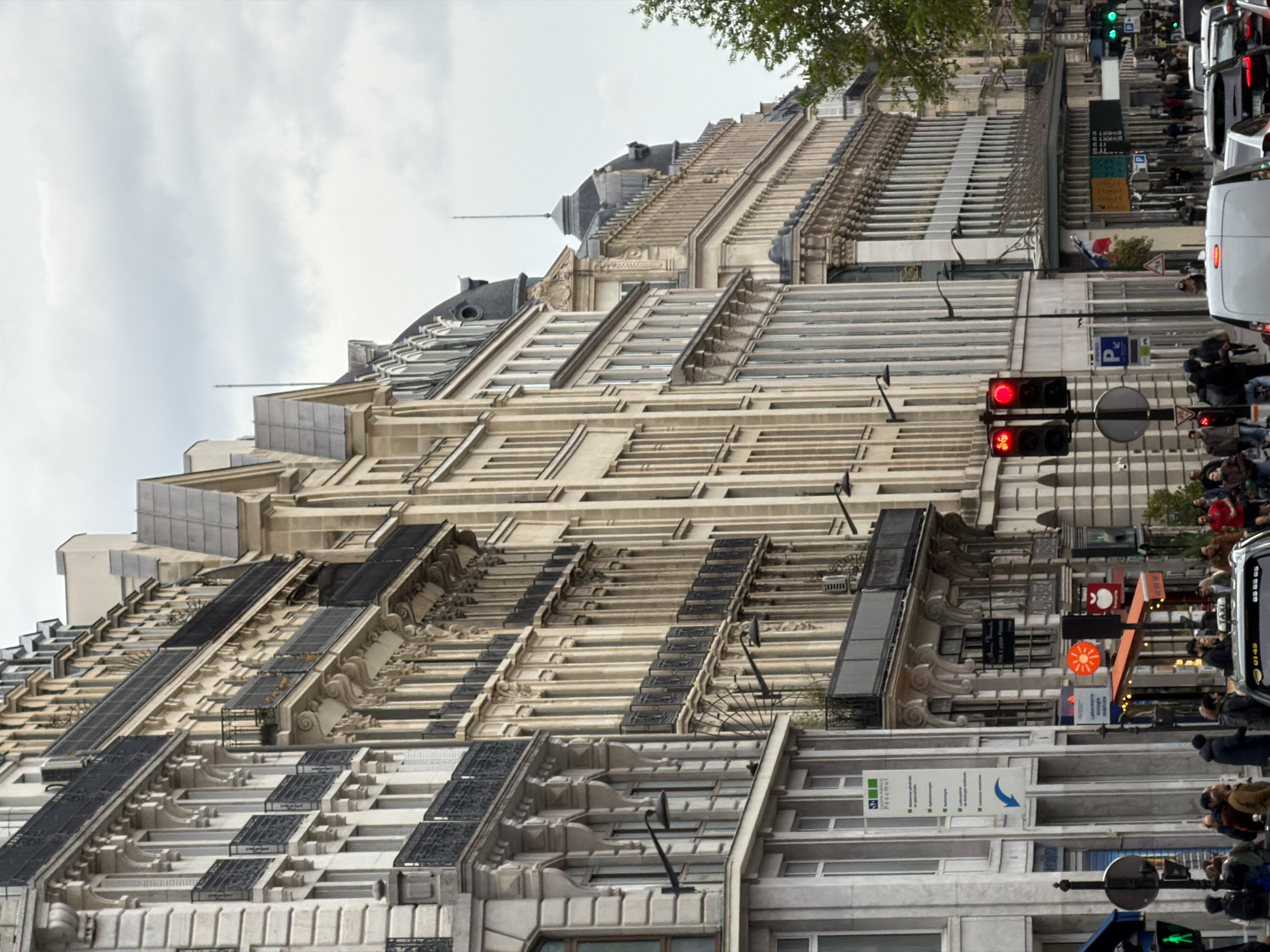 The image depicts a street in Paris, France. A tall, ornate building with multiple stories dominates the left side of the frame. The building features architectural details such as balconies, arched windows, and decorative moldings. On the right side of the frame, a street scene unfolds with pedestrians, vehicles, and traffic lights. The traffic lights show both red and green signals. Various signs and street furniture are visible along the street. The sky is overcast with clouds.