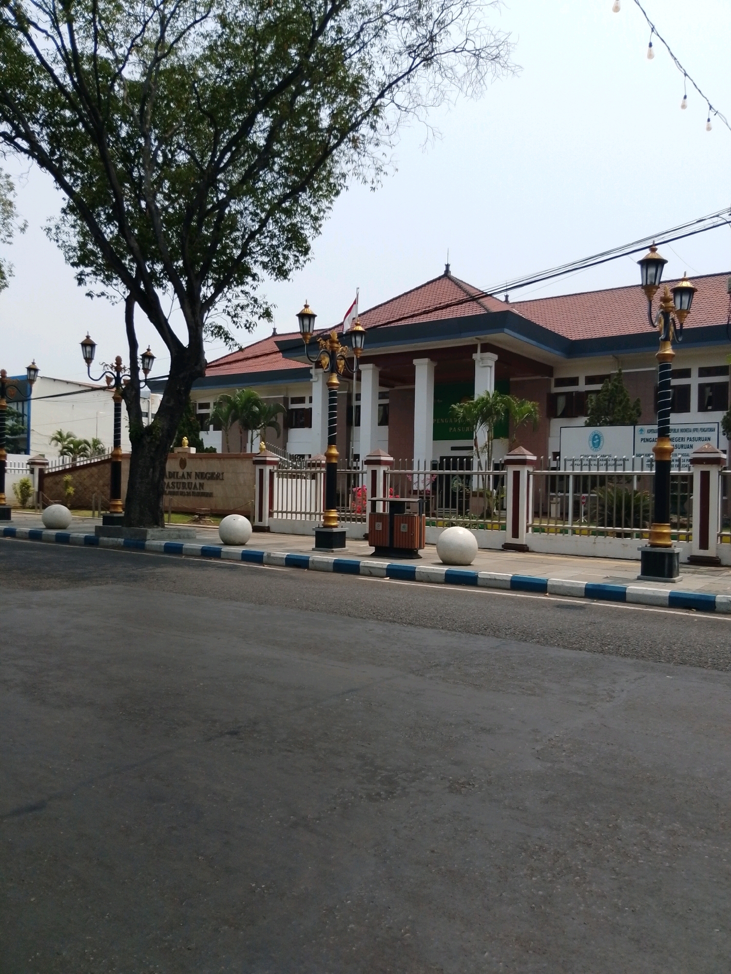 The image shows the facade of the Pengadilan Negeri Pasuruan, a court of law, located in Pasuruan, Indonesia. The building has a red tile roof and white pillars. In front of the building, there is a fence, lampposts, and a sign that reads "Pengadilan Negeri Pasuruan." A large tree is visible to the left of the building. The street in front of the court is paved and has a blue and white striped border. There are no people visible in the image. The sky is overcast.