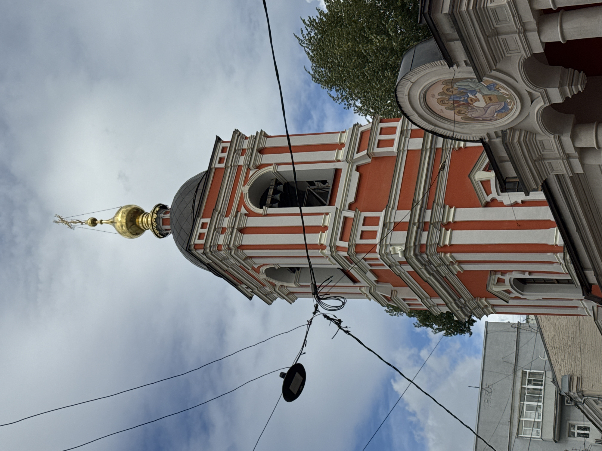 The image shows the bell tower of a red and white Orthodox church in Moscow, Russia. The bell tower is tall and slender, with a golden dome topped with a cross. There are bells visible within the arched openings of the tower. In front of the church, a mosaic depicting three figures is visible above arched colonnades. The sky is cloudy and overcast. Electrical wires and a street lamp are in the foreground, obscuring parts of the church and the sky. A grey apartment building is visible to the left of the church. The overall impression is a view of historic religious architecture within a modern urban environment.