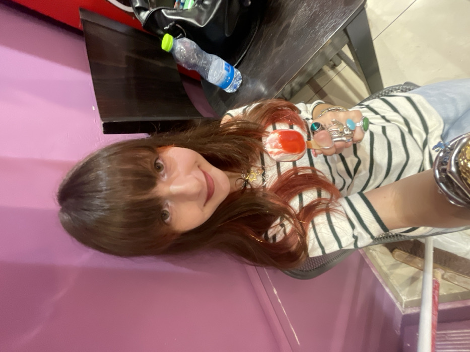 The image captures a cheerful, indoor scene featuring a person smiling at the camera. The image is rotated 90 degrees counter-clockwise, showing the person horizontally, but should be viewed vertically.
The central figure is a person with long, straight brown hair that has noticeable reddish-orange highlights, especially around the face and ends. They have bangs covering their forehead. Their eyes are open, and they have a gentle, pleasant smile. They are wearing a long-sleeved top with alternating light (possibly white or cream) and dark horizontal stripes. Part of their lower body is visible, dressed in light blue jeans or trousers.
On their right hand, which is held up, they are wearing multiple rings, some featuring large, colorful stones, including green and light blue. This hand is holding a clear glass or cup containing a bright red substance, which appears to be a dessert like a sorbet or a drink, with a wooden stick or spoon inserted into it. A necklace with a small, star-shaped pendant (possibly a Star of David) is visible around their neck. On their left wrist, a wide, metallic bracelet is visible.
The background to the left of the person features a solid, purplish-pink wall. To their right, behind and slightly above them, is a dark, possibly wooden or metallic, table or counter. On this surface, a black bag and a clear plastic water bottle with a light green cap are placed. The setting appears to be indoors, possibly in a casual environment like a cafe, salon, or personal space. The lighting is bright and even. No text is visible in the image.