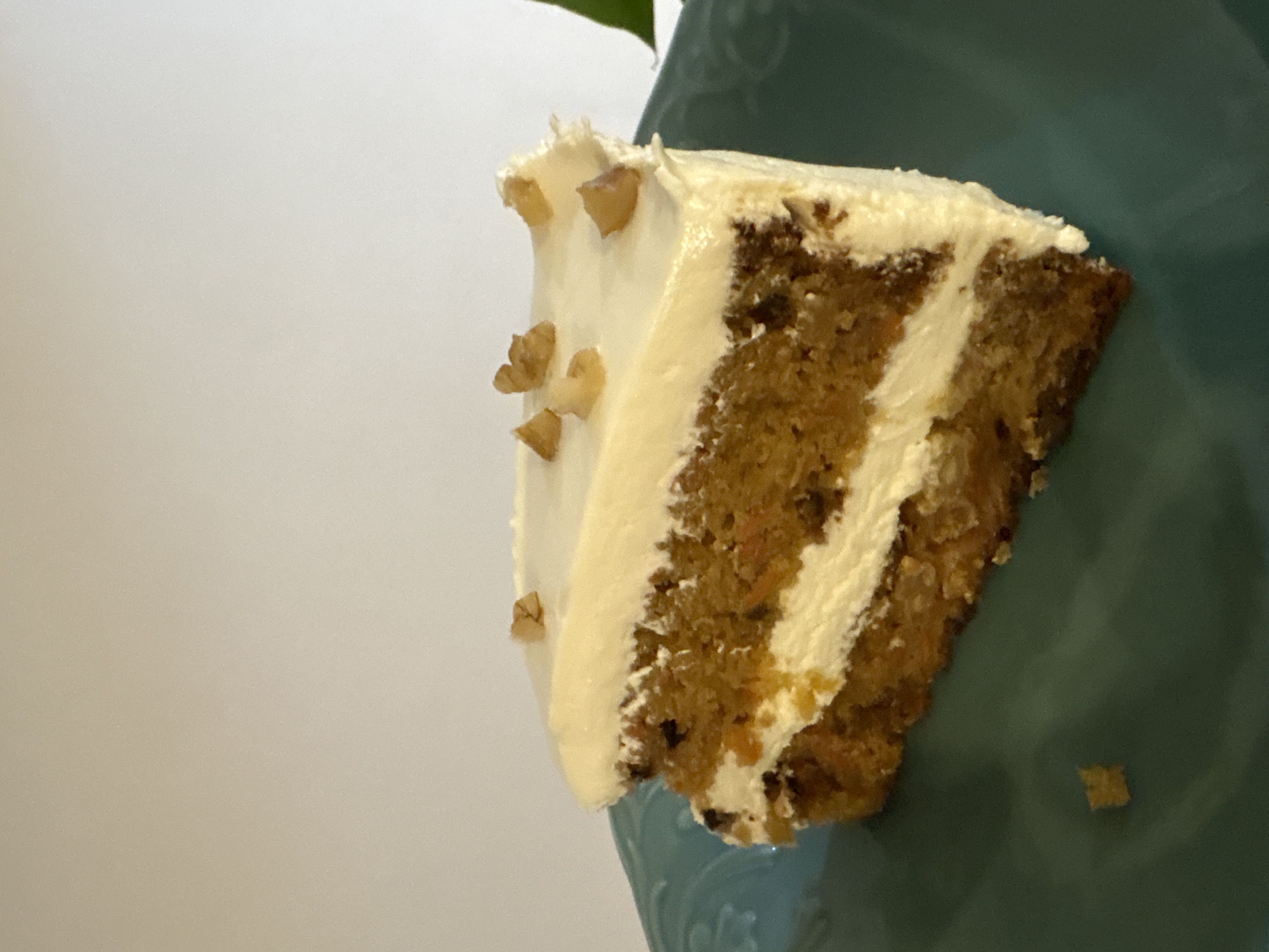 A slice of carrot cake sits on a blue plate. The cake has two layers with cream cheese frosting between and on top, garnished with chopped nuts. The plate is set against a plain white background.