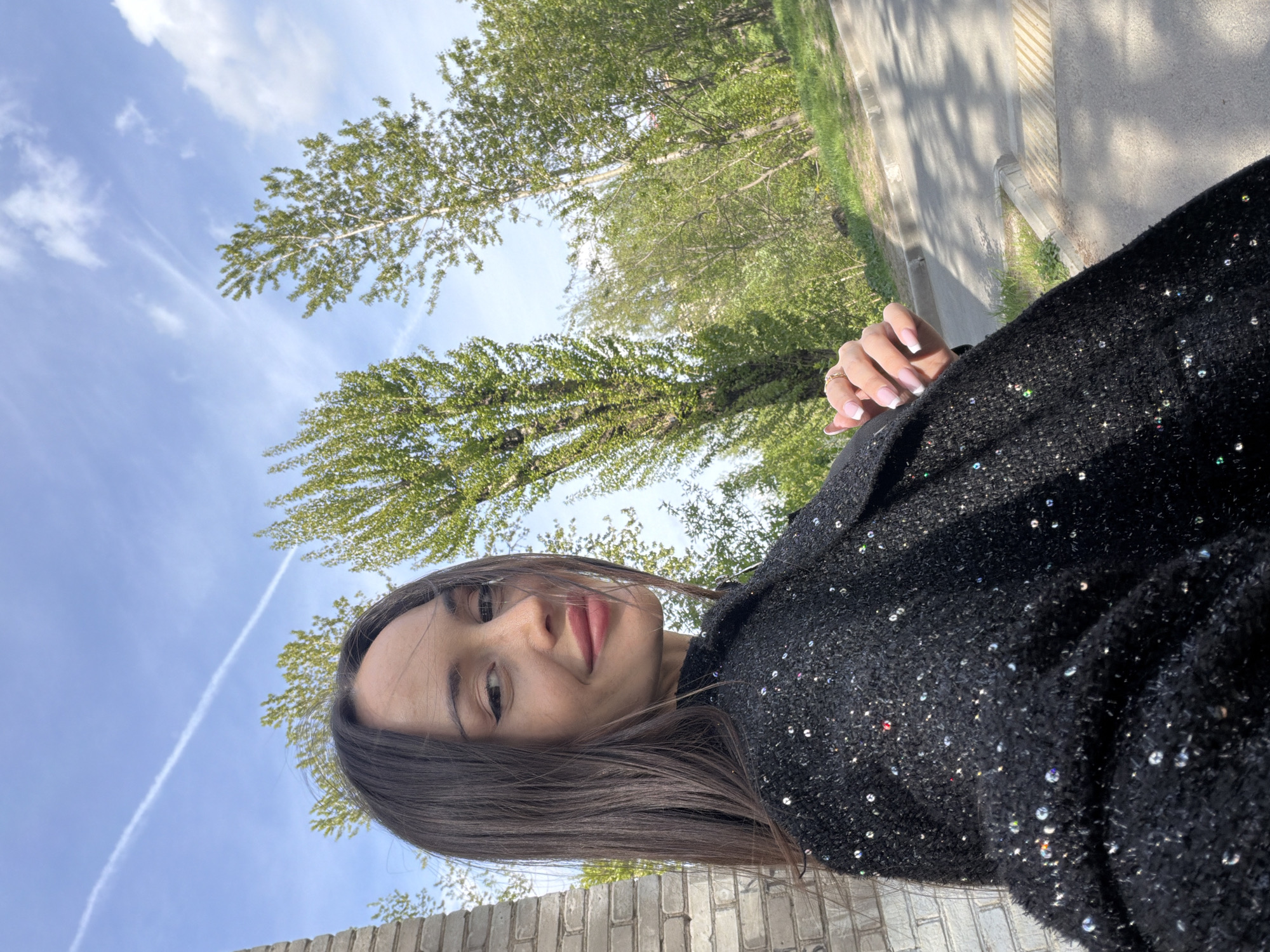 A selfie image features a woman with long brown hair, fair skin, and light pink lips, wearing a black jacket adorned with sparkling sequins. The image is rotated counter-clockwise. She is looking at the camera with a slight smile. Above her is a blue sky with streaky white clouds and the tops of green trees. In the background, there is a gray paved road and a low brick wall. The location is likely Saint Petersburg, Russia, given the architectural style of the brick wall and the general ambiance of the image.