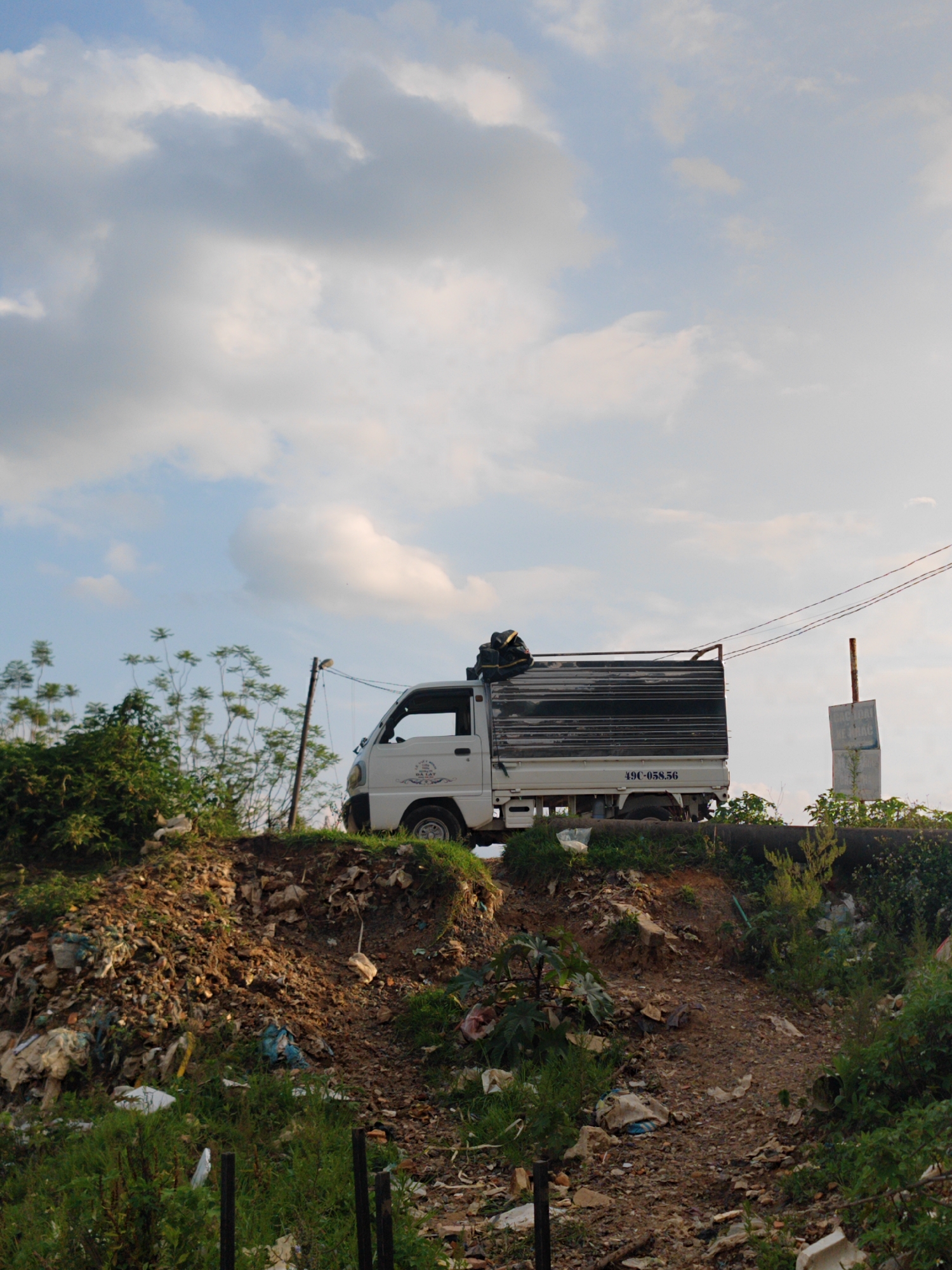 A white truck with a dark, flatbed rear is parked on top of an embankment in what appears to be Cam Ly - Da Lat, Vietnam. The embankment is composed of dirt, trash, and sparse vegetation, suggesting a roadside or unofficial dumping ground. In the foreground is a dirt path leading up to the truck's location, strewn with debris and interspersed with green plants. Behind the truck, more vegetation and a utility pole are visible. A signpost stands on the embankment to the right of the truck, partially obscured. Power lines stretch across the sky. The sky above is a mix of blue and white clouds.
