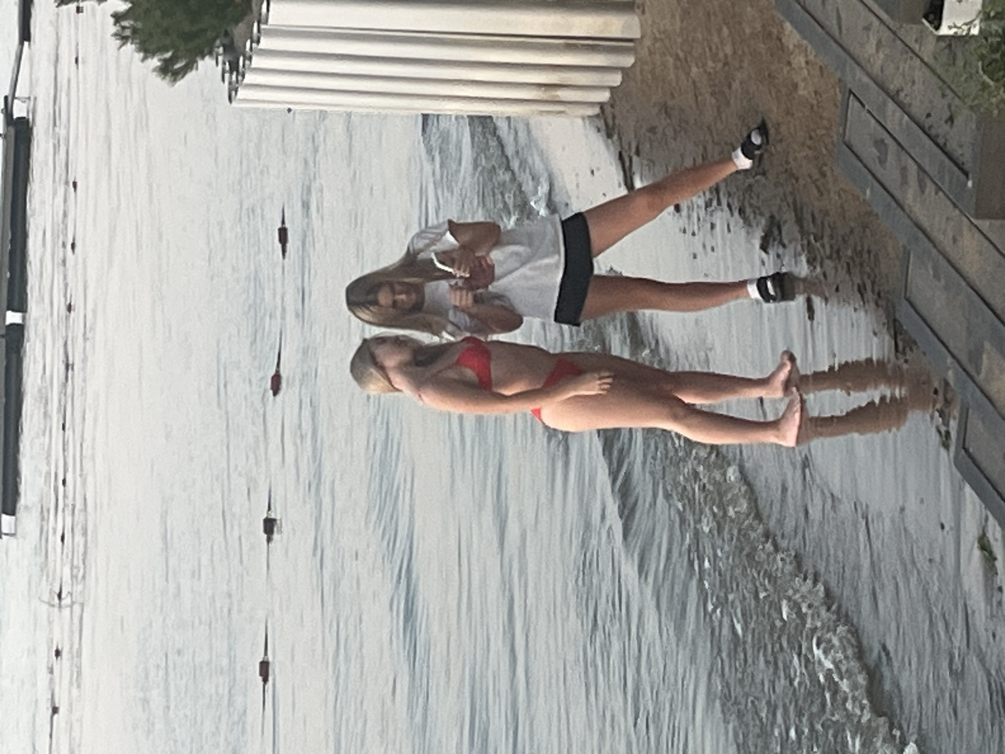 The image depicts two young women standing on the shoreline of Lake Shumeika in Shumeika, Russia. The woman on the left is wearing a red bikini, while the woman on the right is dressed in a white t-shirt and a black skirt. Both are facing each other, with the woman on the right holding a mobile phone. The water is calm, with gentle waves lapping at the shore. In the background, there are buoys on the water and a white, ribbed structure on the beach. The lighting suggests it is either early morning or late evening.