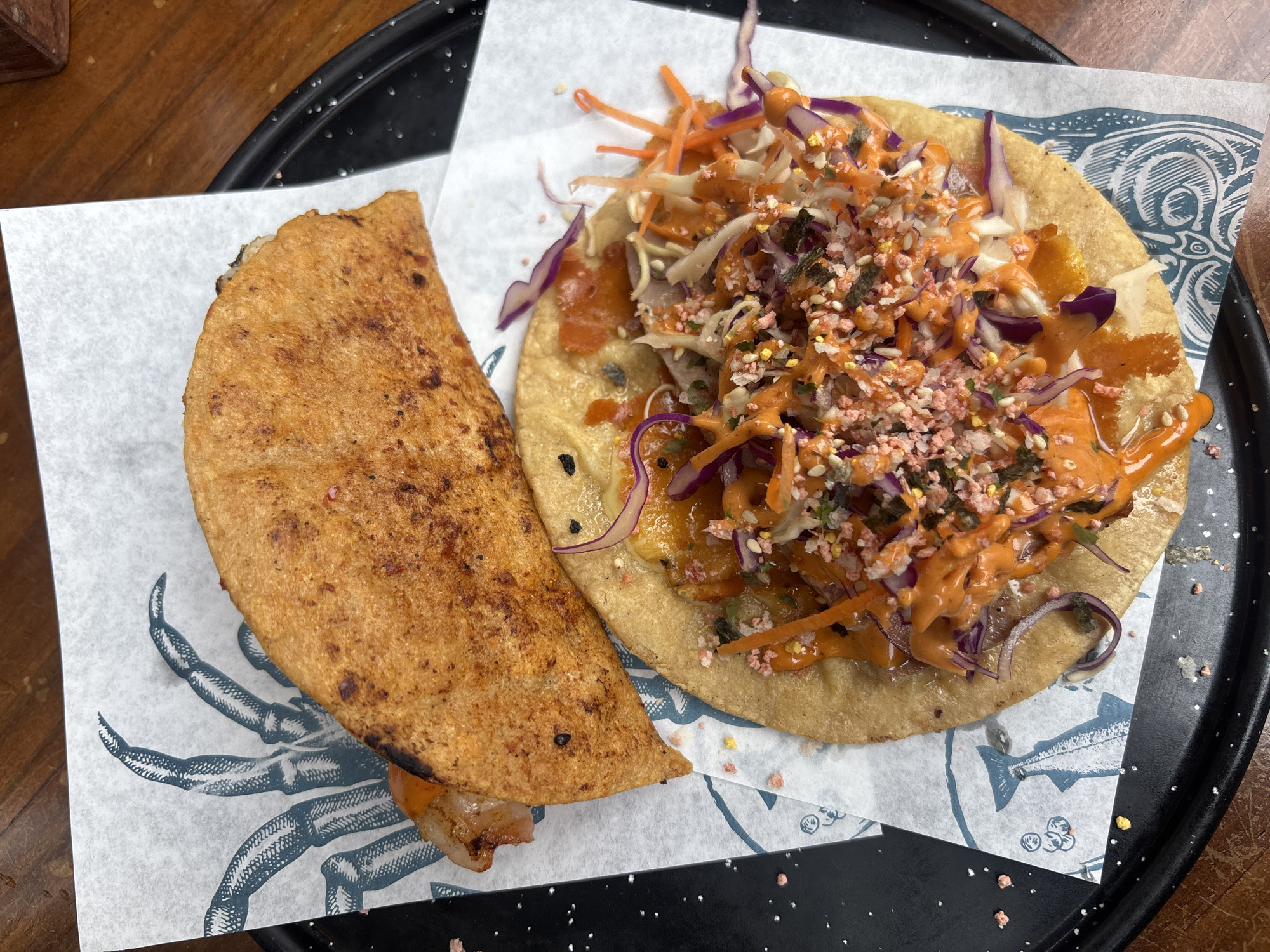 The image shows two tacos on a white paper liner with a seafood design, placed on a black serving plate with white specks. One taco is folded, with a crispy golden-brown shell. The other taco is open-faced, topped with shredded cabbage, carrots, red cabbage strands, and a reddish-orange sauce and pink seasonings. The scene likely takes place in Puebla, Mexico, given the prevalence of tacos in Mexican cuisine.