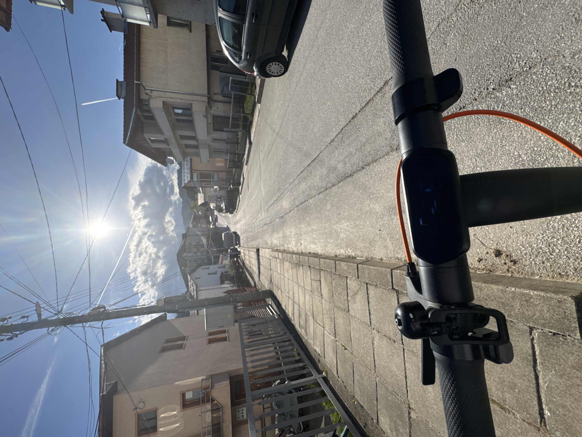 A sunny day in Borino, Bulgaria, captured from the perspective of someone riding an electric scooter down a quiet street. The scooter's handlebars and control panel are in the foreground. The street extends into the distance, flanked by buildings of varying heights and architectural styles, typical of a small European town. A parked car is visible on the right side of the street, partially cut off by the image frame. Power lines crisscross the sky, contrasting with the blue sky and scattered clouds. A contrail from a high-flying aircraft is visible in the upper left corner. The sun shines brightly, creating strong shadows on the street and buildings. The overall impression is one of a peaceful residential area on a clear, bright day.